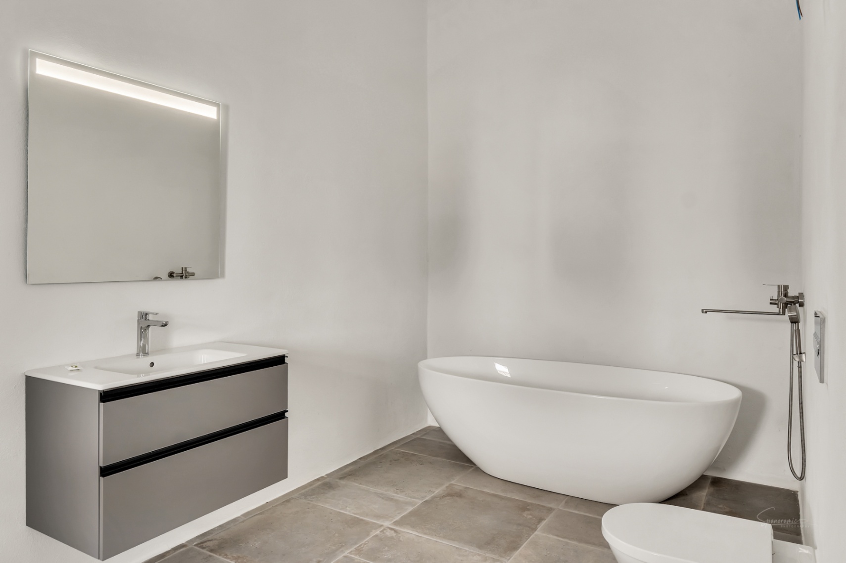 Baño moderno con bañera exenta ovalada, mueble suspendido en gris con lavabo integrado y espejo con iluminación LED sobre suelos de piedra natural.