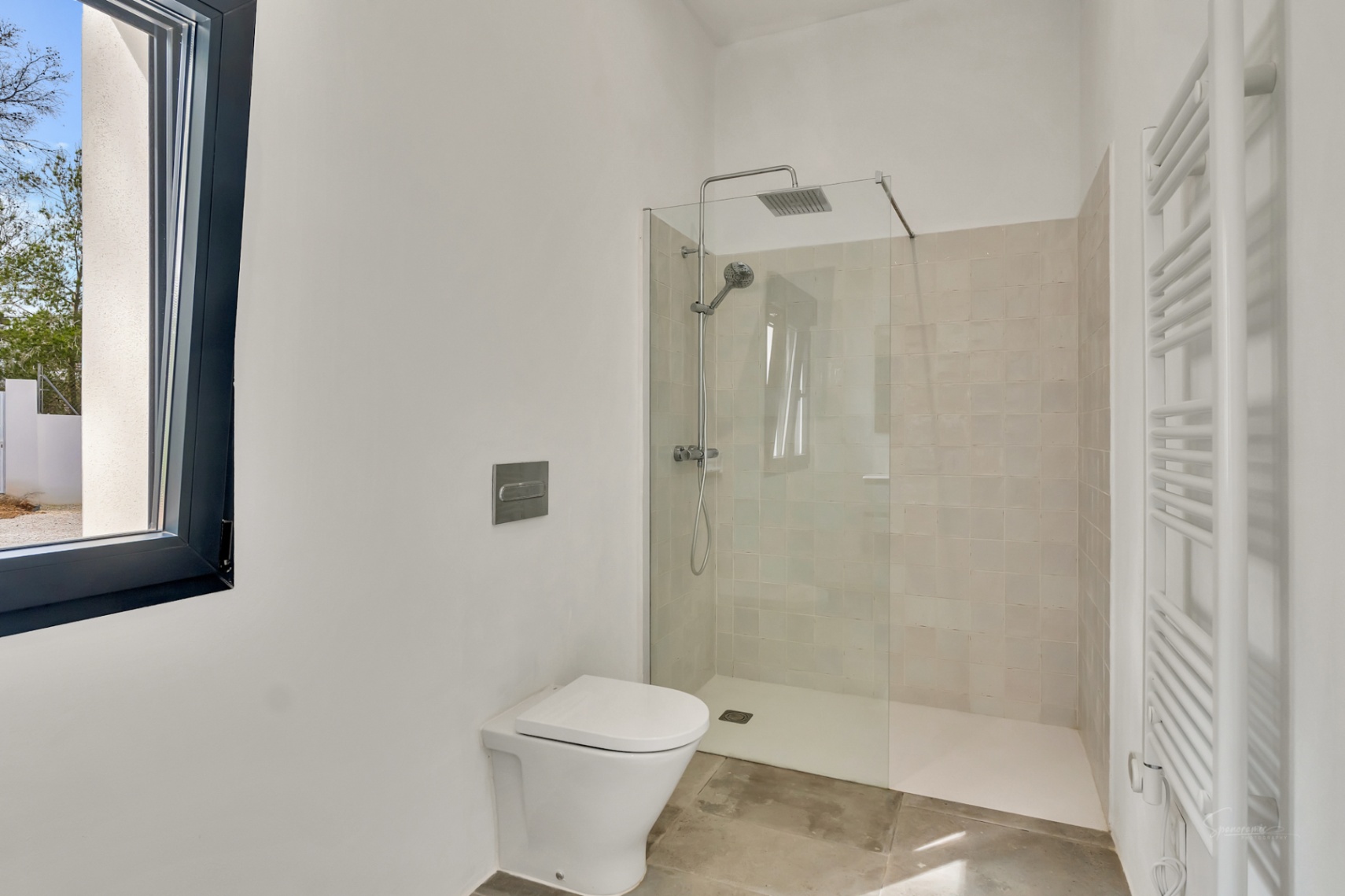 Baño moderno con ducha a ras de suelo, mampara de cristal y azulejos neutros. Dispone de inodoro suspendido, radiador toallero blanco y suelos de piedra natural.