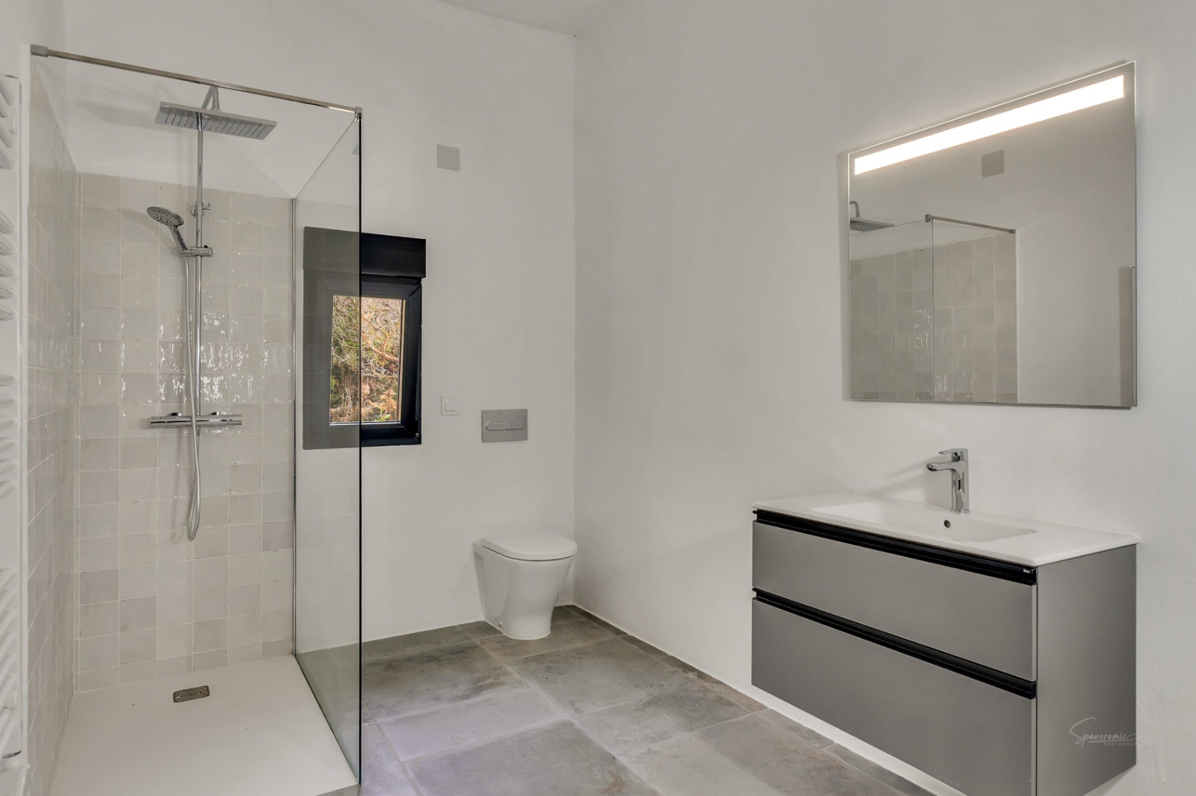 Baño moderno con ducha a ras de suelo, mampara de cristal y grifería de efecto lluvia. Incluye un mueble suspendido gris y espejo con iluminación integrada sobre suelo de piedra.