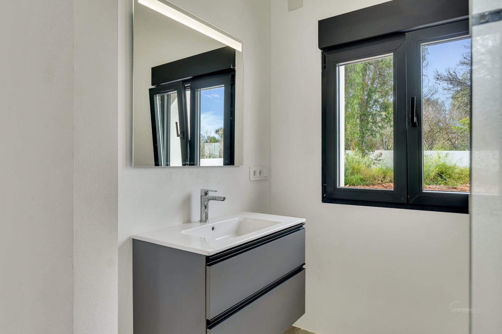 Baño moderno con mueble suspendido en gris antracita, lavabo blanco y espejo con iluminación LED. La ventana de carpintería negra ofrece luz natural y vistas al jardín.