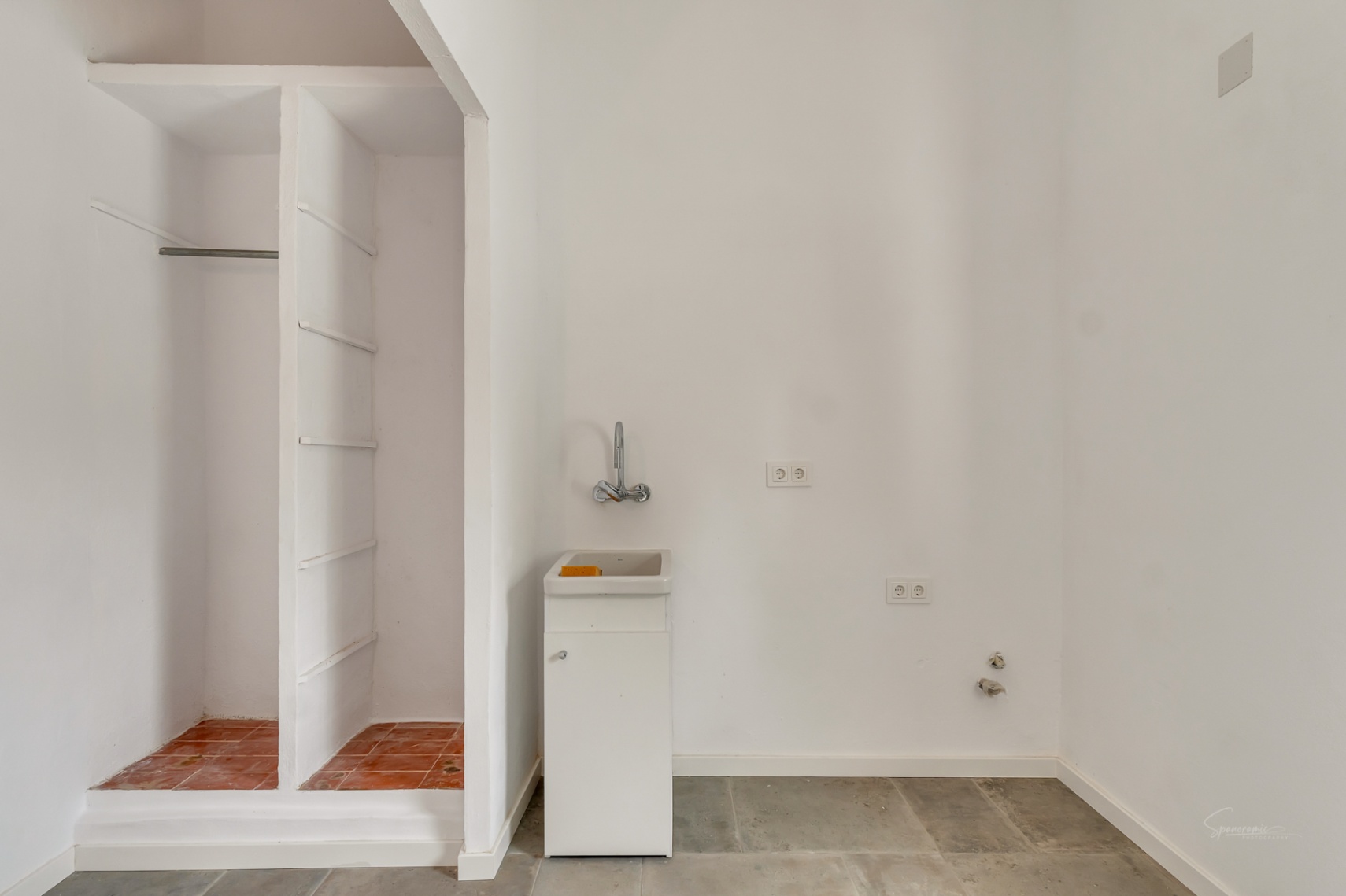 Cuarto de servicio funcional con lavabo compacto, tomas eléctricas y un nicho de almacenamiento de obra con base de terracota y suelos de piedra.