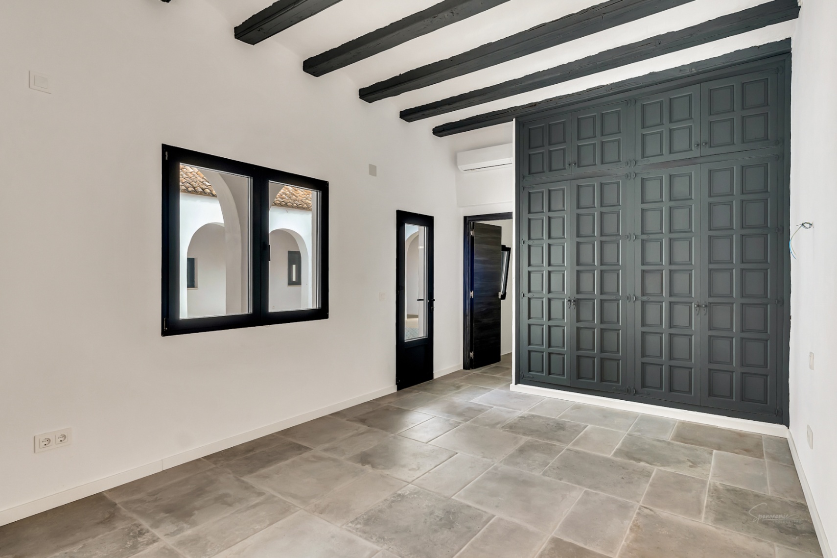 Dormitorio con vigas de madera oscura, suelos de piedra natural y un gran armario empotrado. El ventanal ofrece vistas directas al patio interior de estilo mediterráneo con arcos.