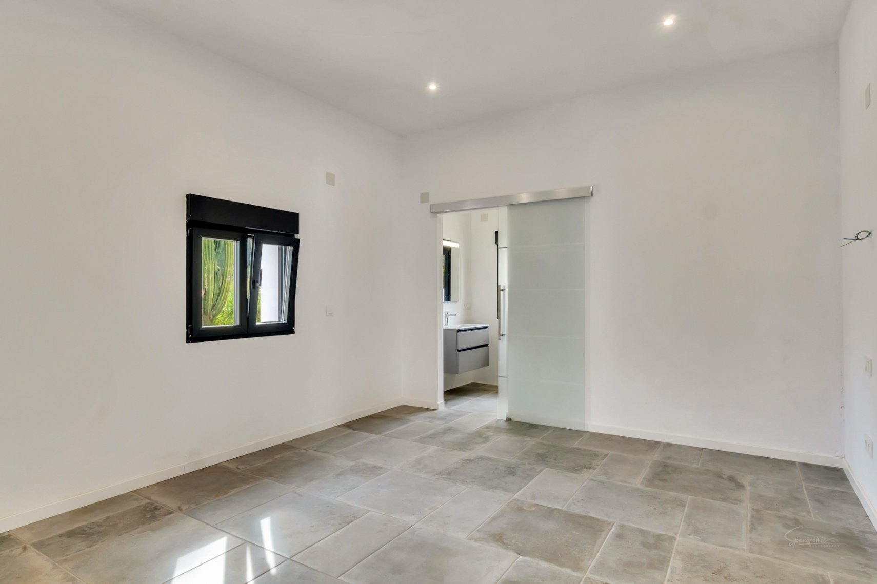 Dormitorio luminoso con suelos de piedra natural, ventana de carpintería negra y puerta corredera de cristal translúcido que da acceso al baño en suite.