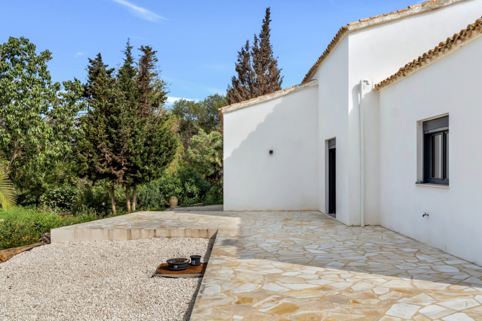 Exterior de la villa con fachada de estuco blanco y tejado de terracota, destacando una amplia terraza de piedra natural rodeada de jardines maduros y zonas de grava.