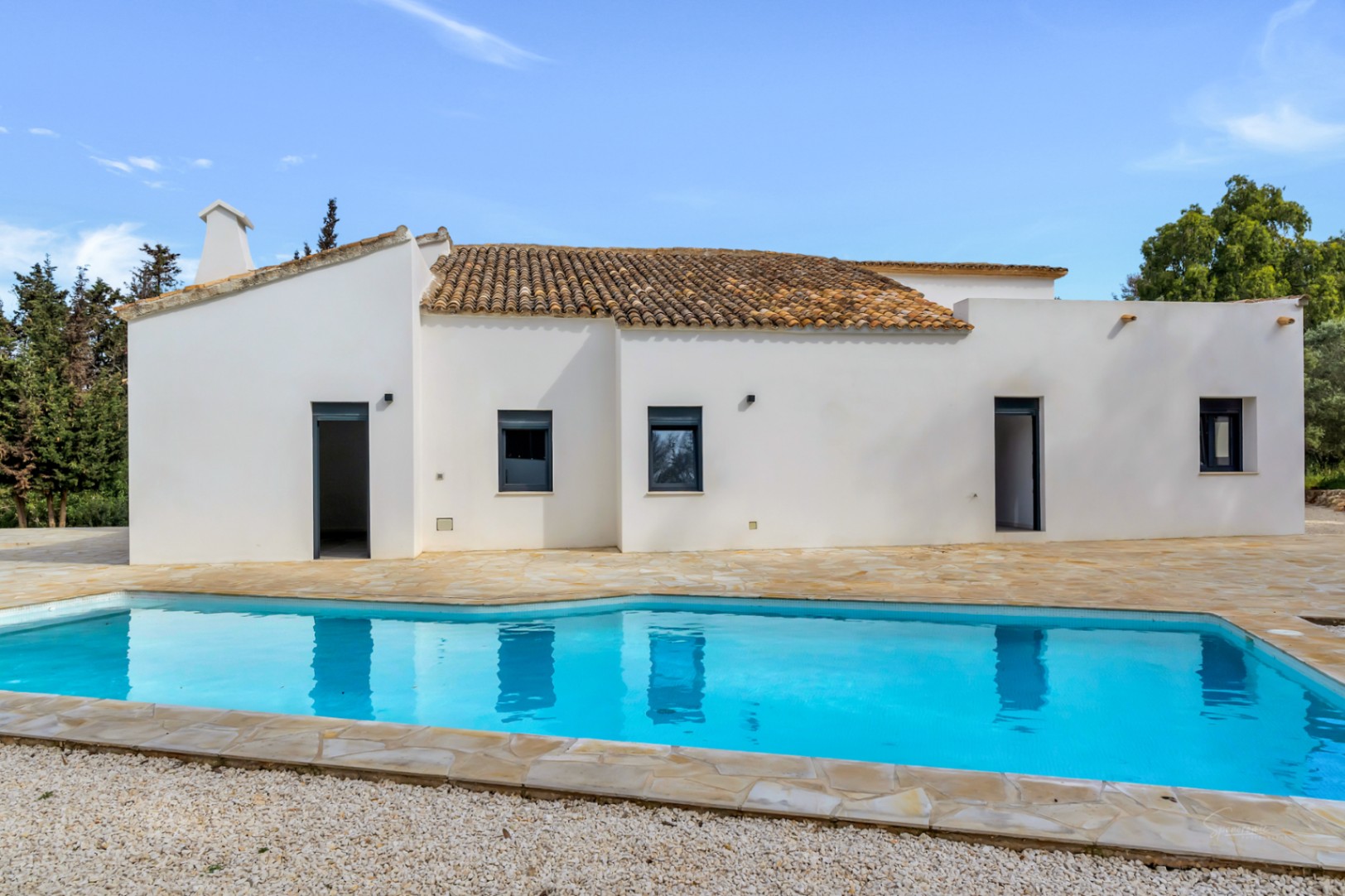 Exterior de la villa con piscina privada y amplia terraza de piedra natural. La fachada blanca resalta con carpintería oscura y el tejado de teja tradicional mediterránea.