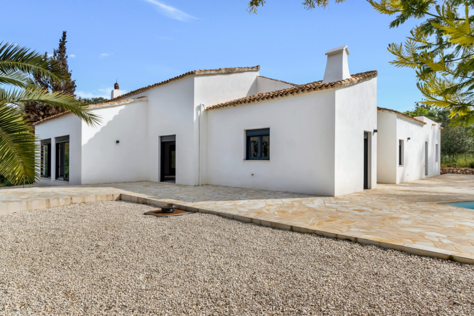 Fachada exterior de villa mediterránea con paredes de estuco blanco, tejado de terracota y amplia terraza de piedra natural junto a zona de grava y jardín.