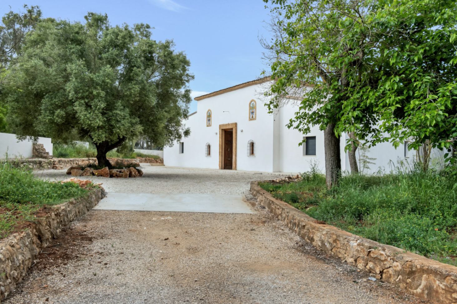 Fachada principal de estilo mediterráneo con muros de estuco blanco, marcos de piedra tosca y entrada flanqueada por árboles maduros en una amplia parcela llana.