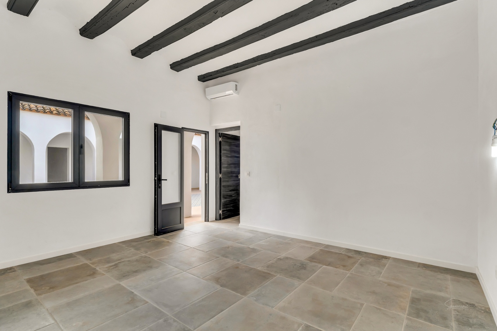 Habitación con vigas de madera oscura expuestas, suelos de piedra natural y vistas al patio interior arqueado a través de carpintería negra.