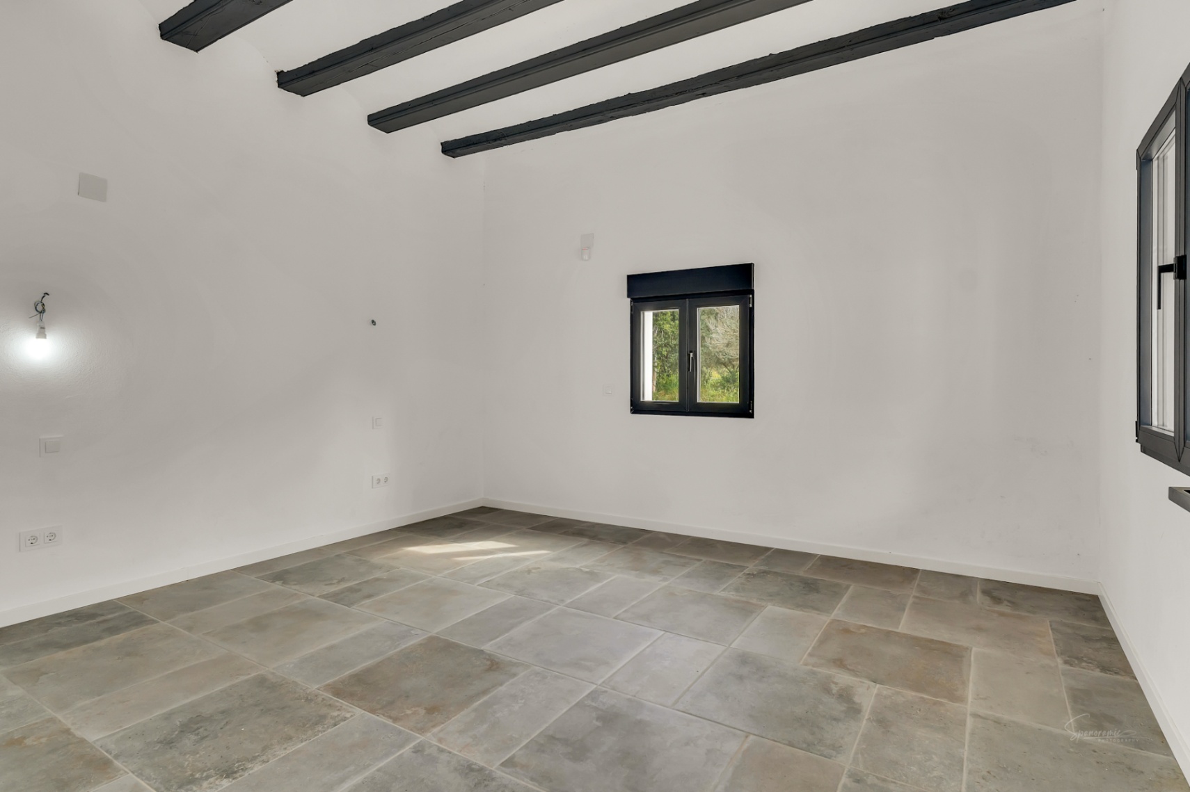 Habitación diáfana con suelos de piedra natural, vigas de madera oscura vistas en el techo y paredes blancas que maximizan la luz natural de las ventanas de carpintería negra.