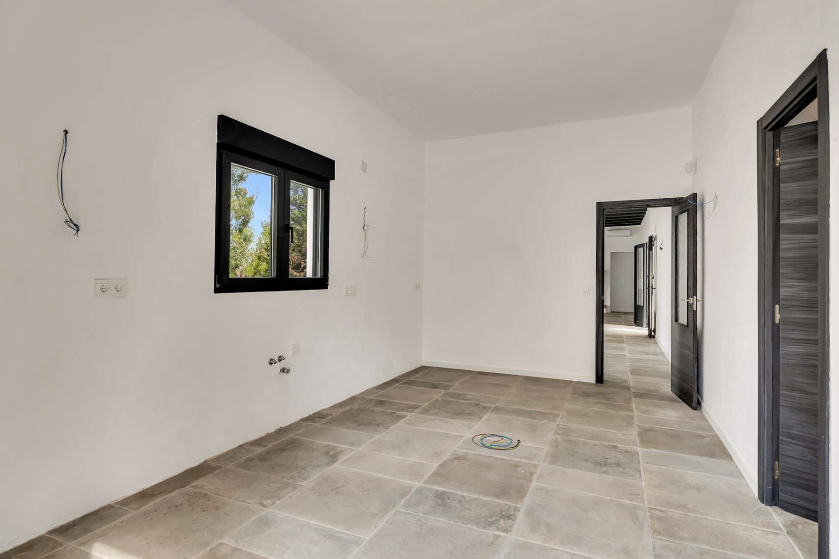 Habitación en proceso de reforma con suelos de gres porcelánico en tonos grises, paredes blancas y carpintería exterior en negro con vistas al jardín.