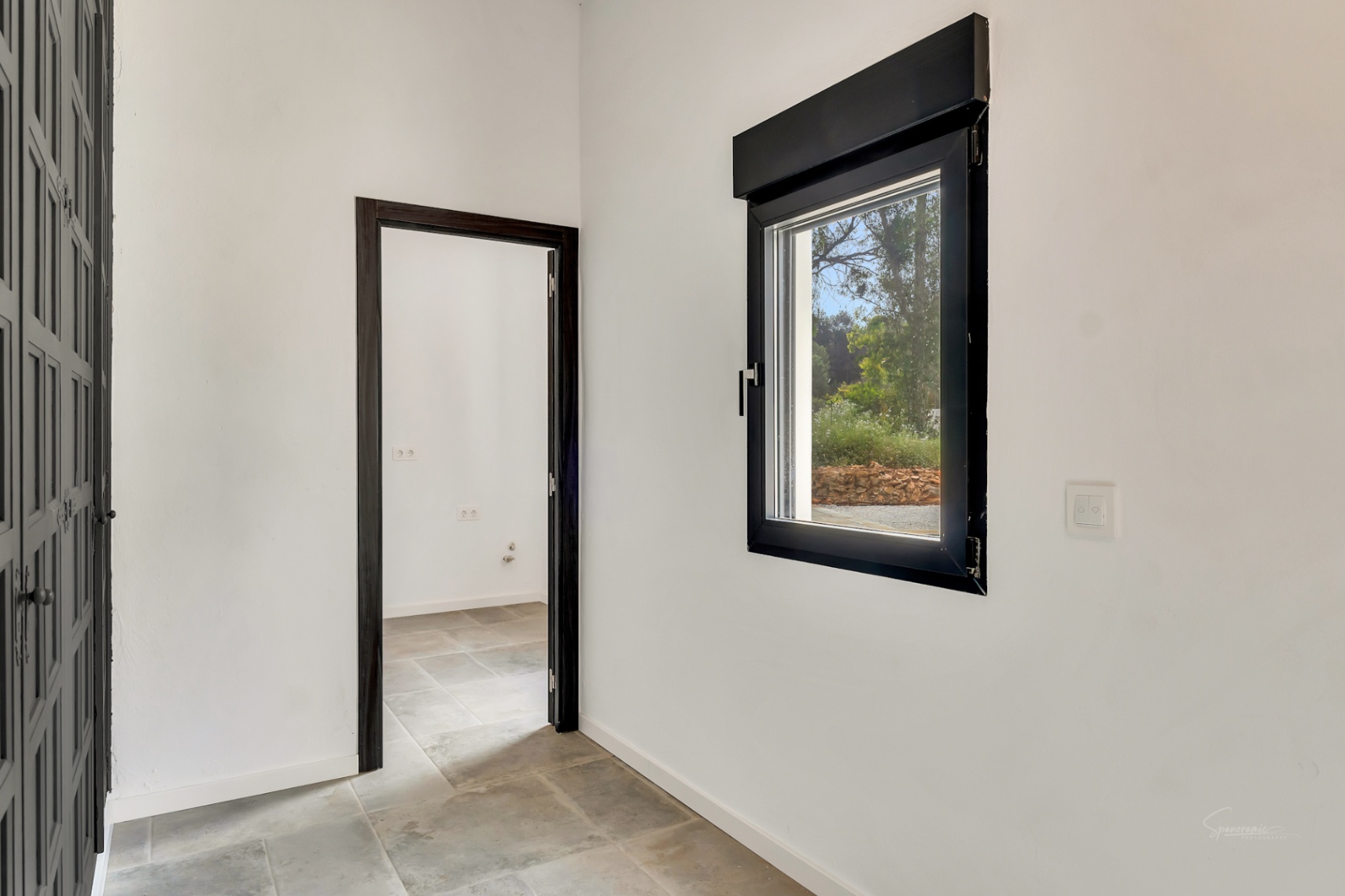 Pasillo con suelos de piedra natural y carpintería en tonos oscuros. La ventana con marco negro permite la entrada de luz natural y ofrece vistas a la zona exterior.