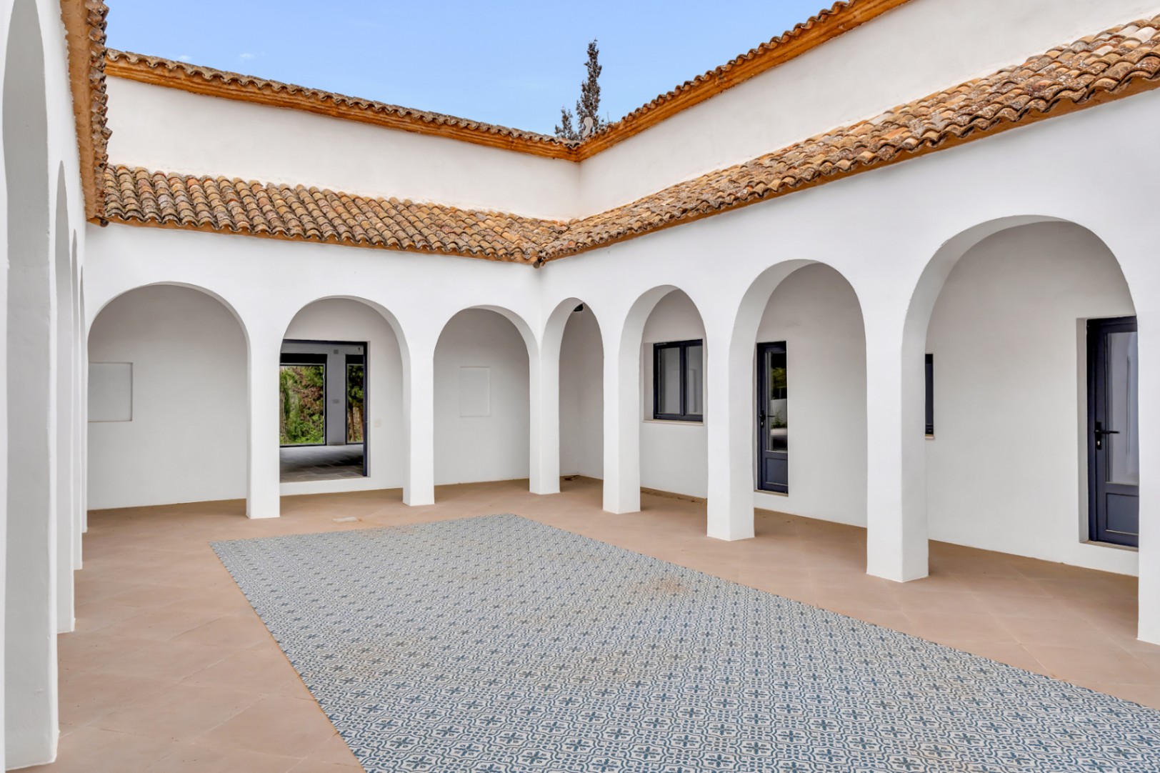 Patio interior central con arquería blanca de estilo mediterráneo, suelos de terracota y un mosaico central de baldosas decorativas bajo un tejado de teja tradicional.
