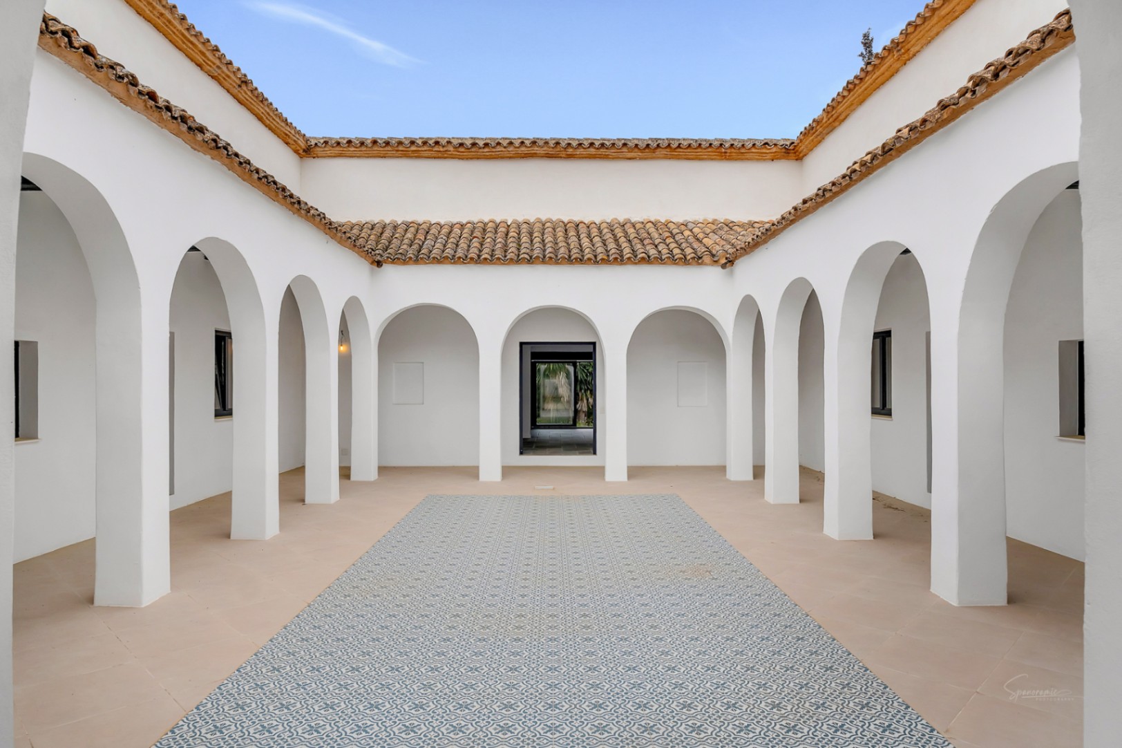 Patio interior central de estilo mediterráneo con arcos blancos simétricos, tejado de terracota y suelo decorado con baldosas hidráulicas estampadas en azul.
