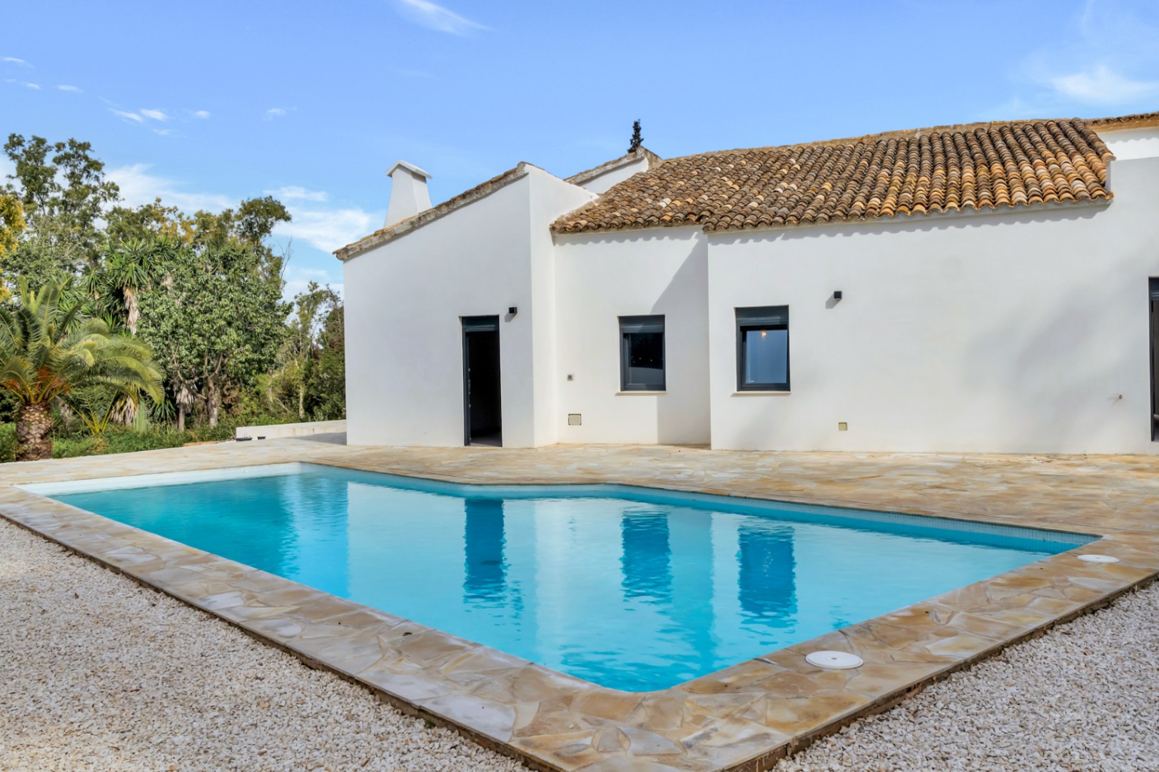 Piscina exterior rodeada de una amplia terraza de piedra natural y grava blanca, junto a una fachada de estilo mediterráneo con tejado de terracota.
