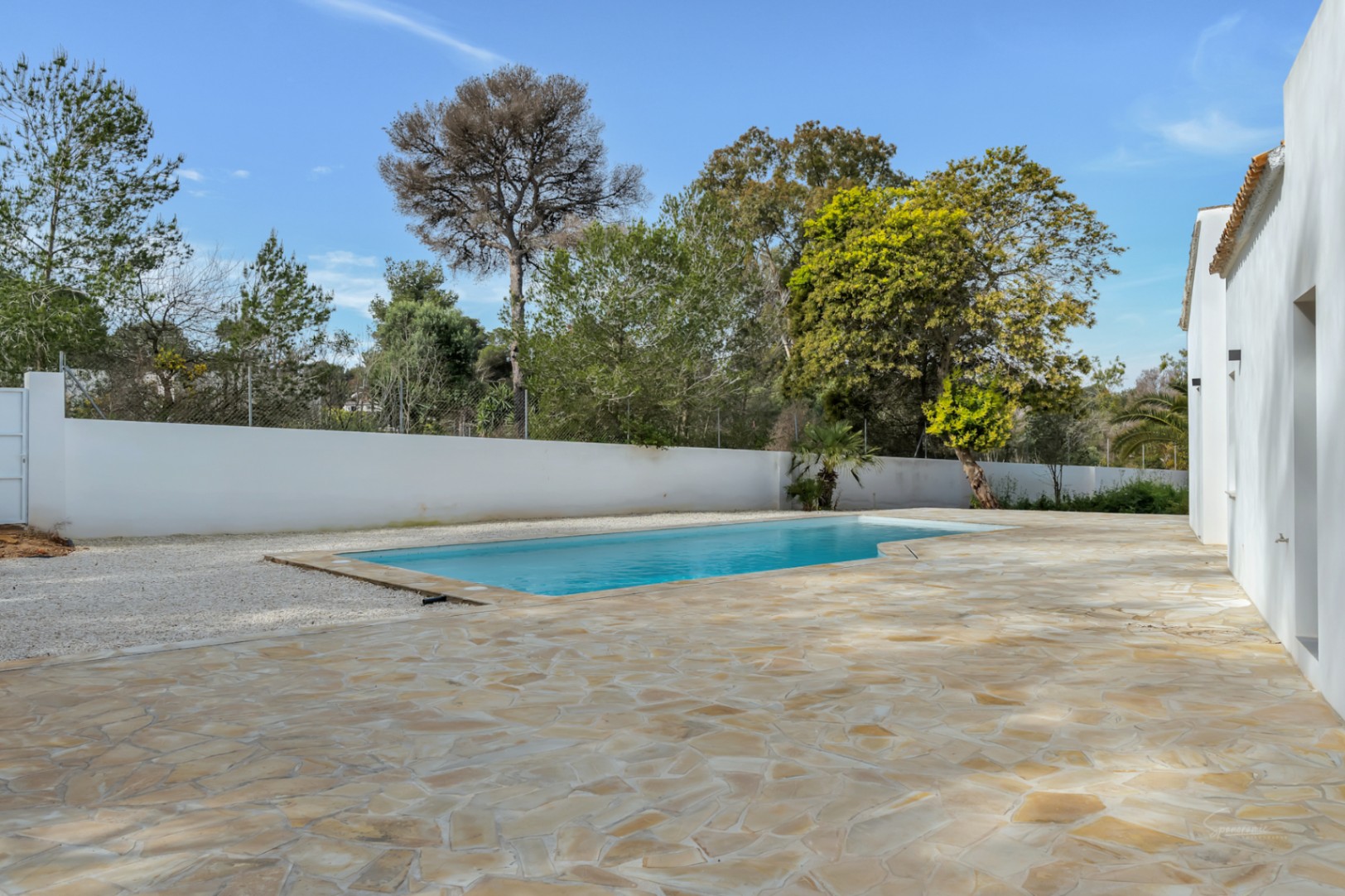 Piscina exterior rodeada de una amplia terraza de piedra natural y muros blancos, integrada en una parcela con vegetación mediterránea madura.