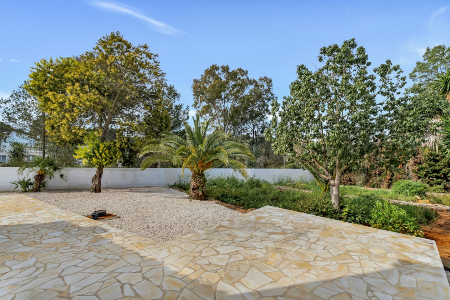 Terraza exterior pavimentada con piedra natural de corte irregular, que da paso a un jardín mediterráneo con palmeras y árboles maduros sobre una zona de grava.