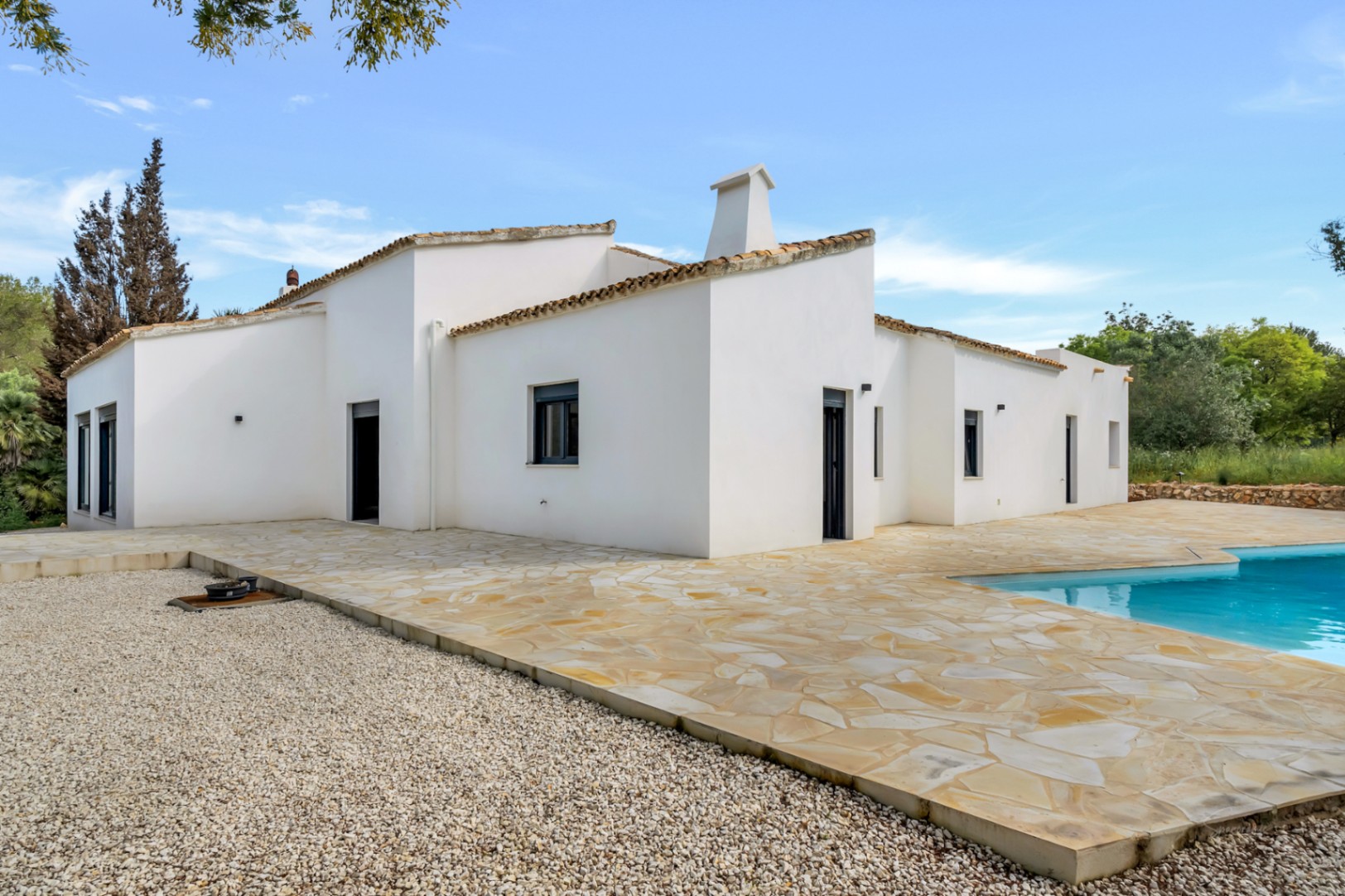 Vista exterior de la villa de una planta con fachada de estuco blanco, tejado de terracota y una amplia terraza de piedra natural junto a la piscina.