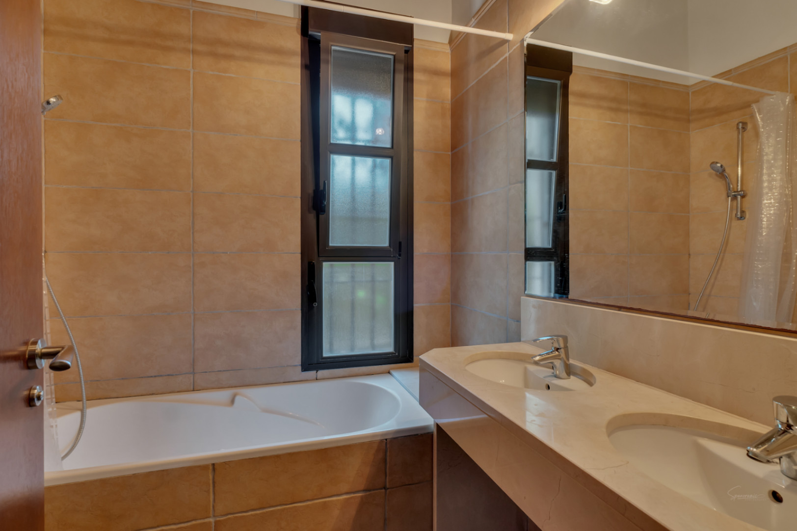 Baño con encimera de piedra de doble seno, bañera integrada y revestimiento de azulejos en tonos tierra. Incluye una ventana vertical que aporta luz natural tamizada.