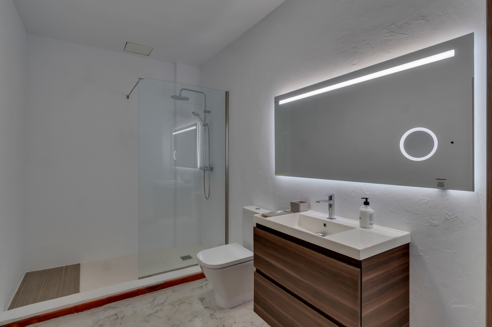 Baño moderno con mueble de madera oscura, lavabo integrado y espejo retroiluminado. Incluye una ducha de obra con mampara de cristal y acabados en tonos blancos.