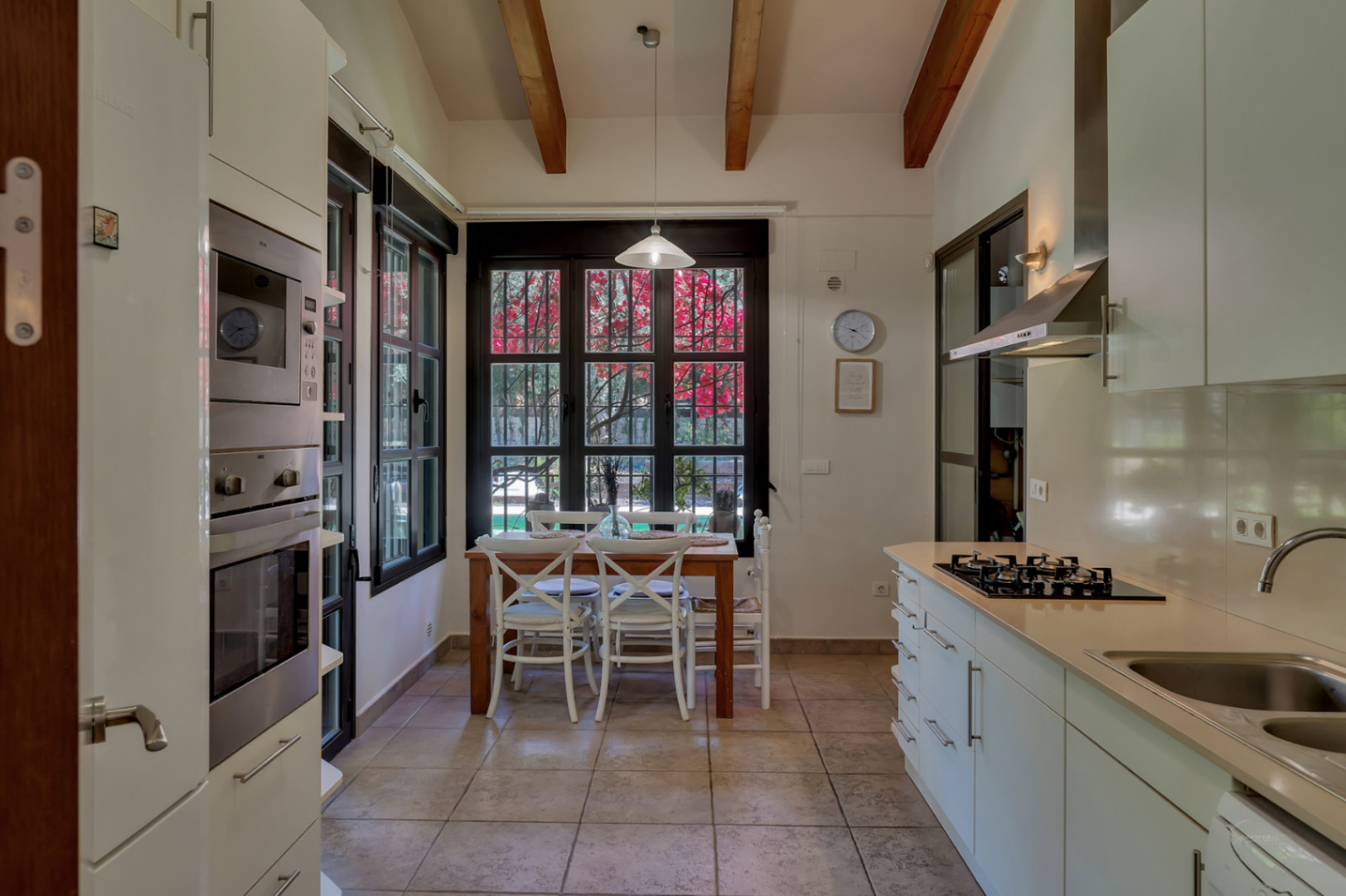 Cocina funcional con vigas de madera vistas, electrodomésticos de acero inoxidable y zona de desayuno junto a ventanales con vistas al jardín mediterráneo.