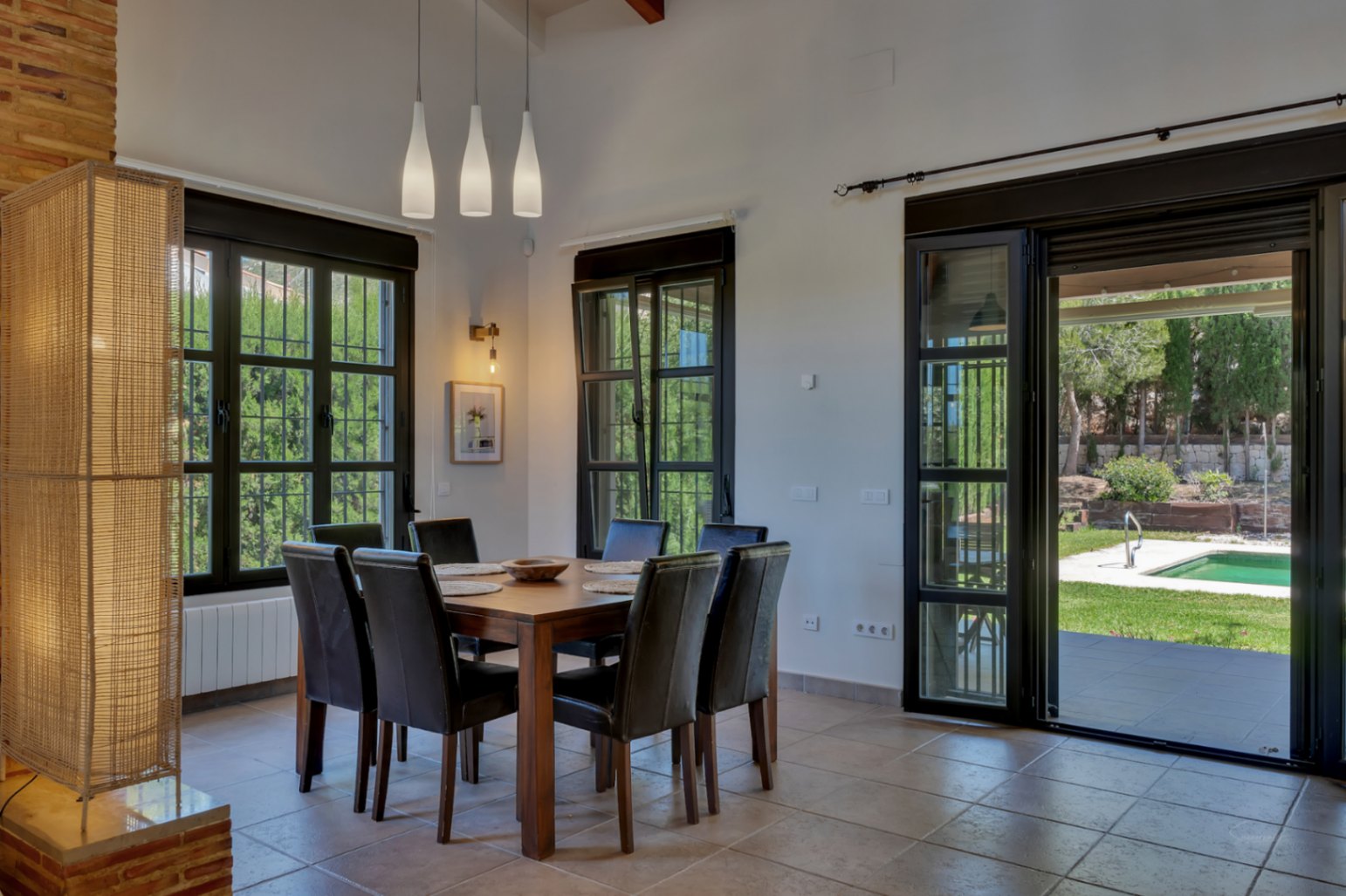 Comedor con mesa de madera, sillas de cuero y lámparas colgantes, conectado al jardín y la piscina mediante amplios ventanales de carpintería negra.