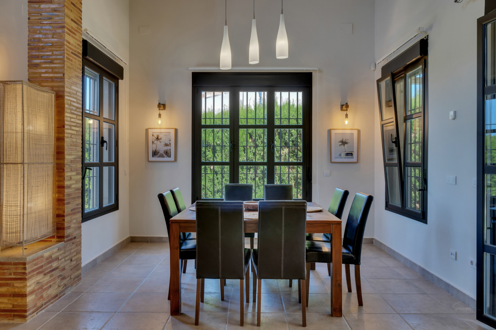 Comedor con mesa de madera y seis sillas de cuero negro, iluminado por lámparas colgantes y apliques de pared junto a ventanales con marcos oscuros y vistas al jardín.