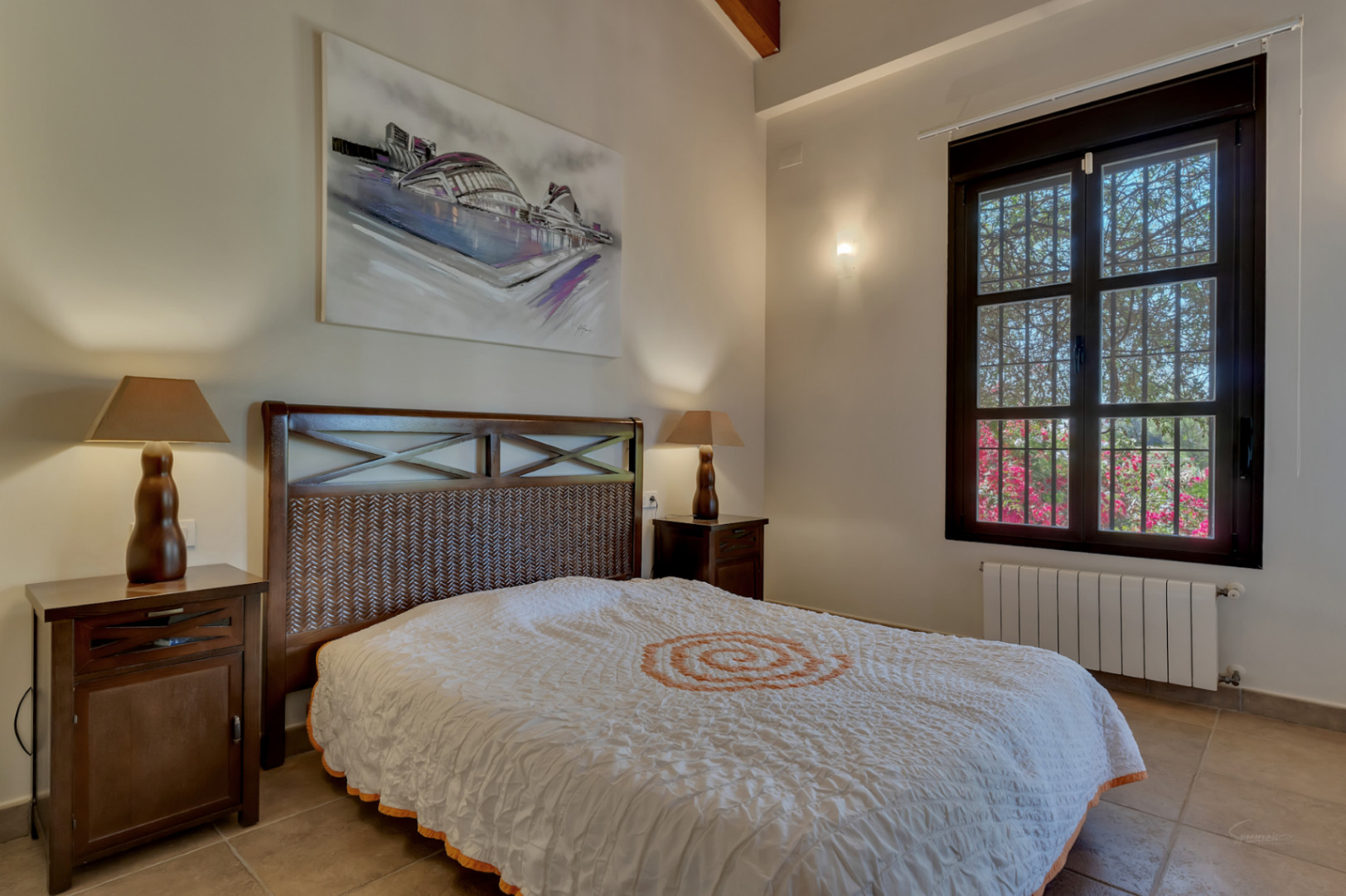 Dormitorio con cama de madera oscura, cabecero trenzado y vigas vistas. Incluye ventanal con marco de madera, suelos cerámicos y vistas al jardín con buganvillas.