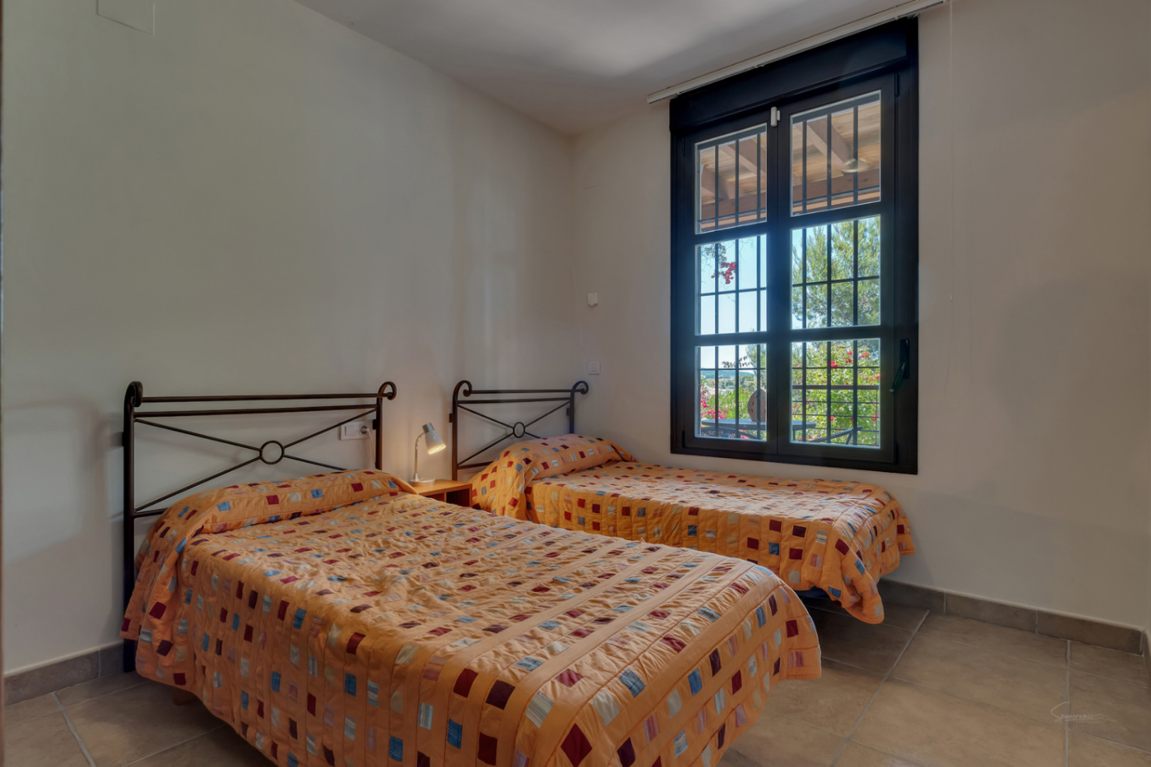 Dormitorio con dos camas individuales, cabeceros de forja y suelos cerámicos. La ventana con marco oscuro ofrece vistas directas al jardín mediterráneo.