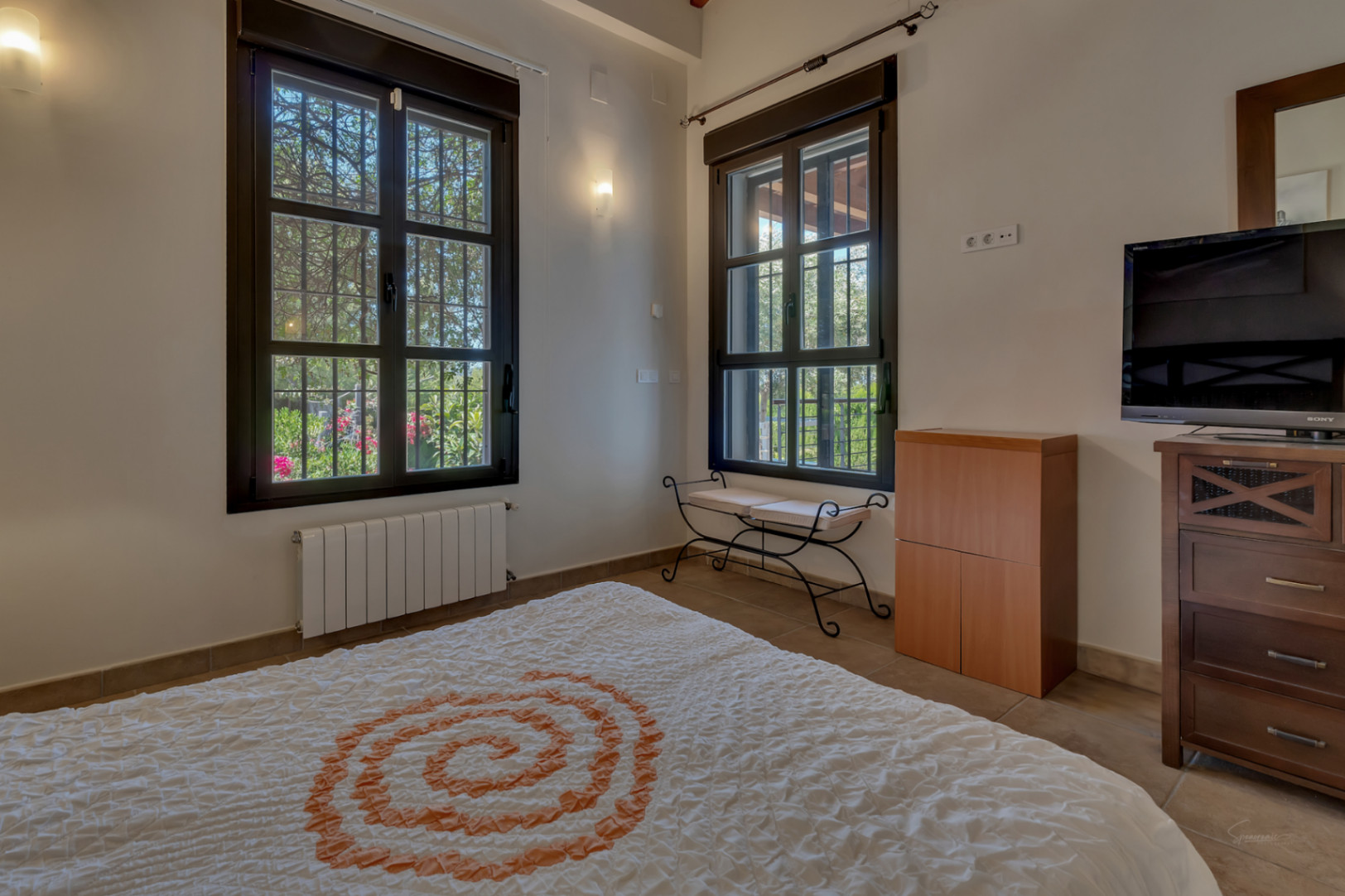 Dormitorio luminoso con amplios ventanales de madera oscura, radiador de pared, mobiliario funcional y vistas directas al jardín mediterráneo.