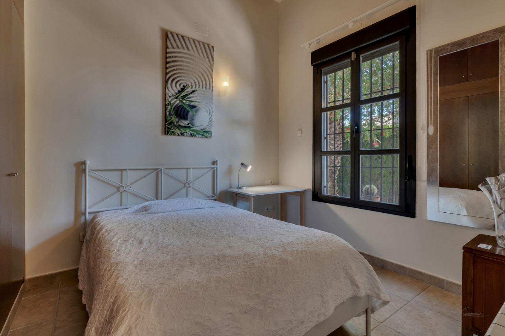 Dormitorio luminoso con cama de forja blanca, escritorio funcional y ventana con marco oscuro que ofrece vistas al jardín mediterráneo.