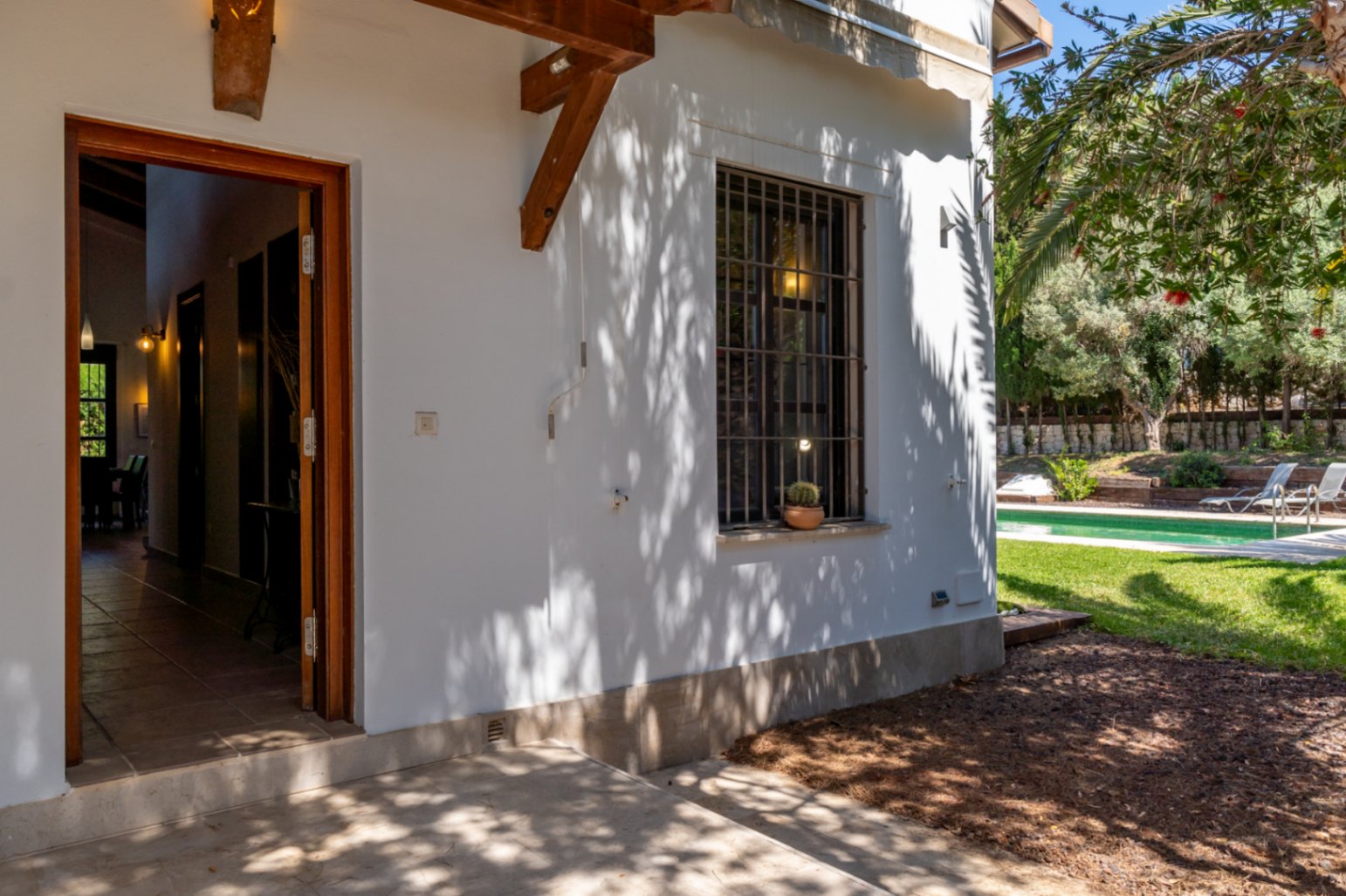 Entrada exterior con puerta de madera abierta hacia el pasillo, fachada blanca con vigas decorativas y vistas al jardín con piscina y césped.
