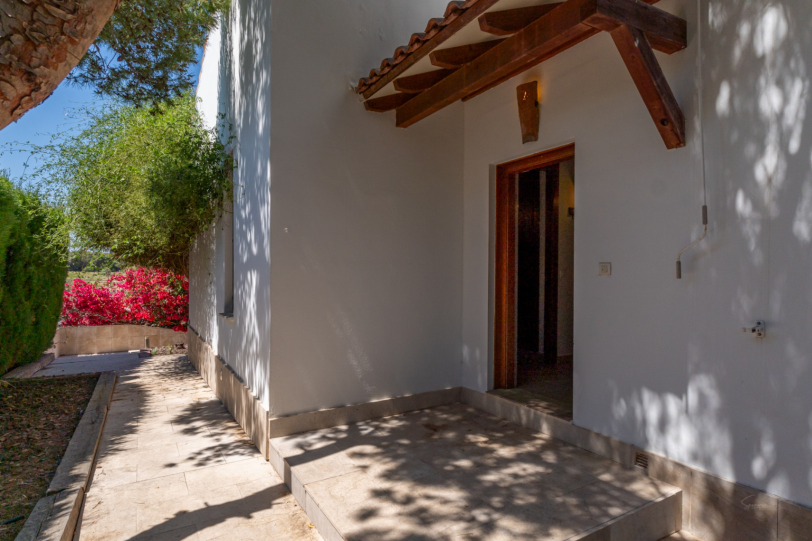 Entrada lateral con porche de vigas de madera y tejas, suelo de piedra natural y vistas al jardín con buganvillas vibrantes.