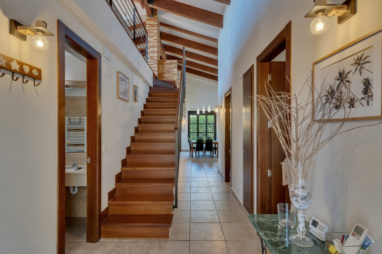 Pasillo distribuidor con techos de vigas de madera vistas, escalera de madera noble y suelos cerámicos que conducen a un luminoso comedor.