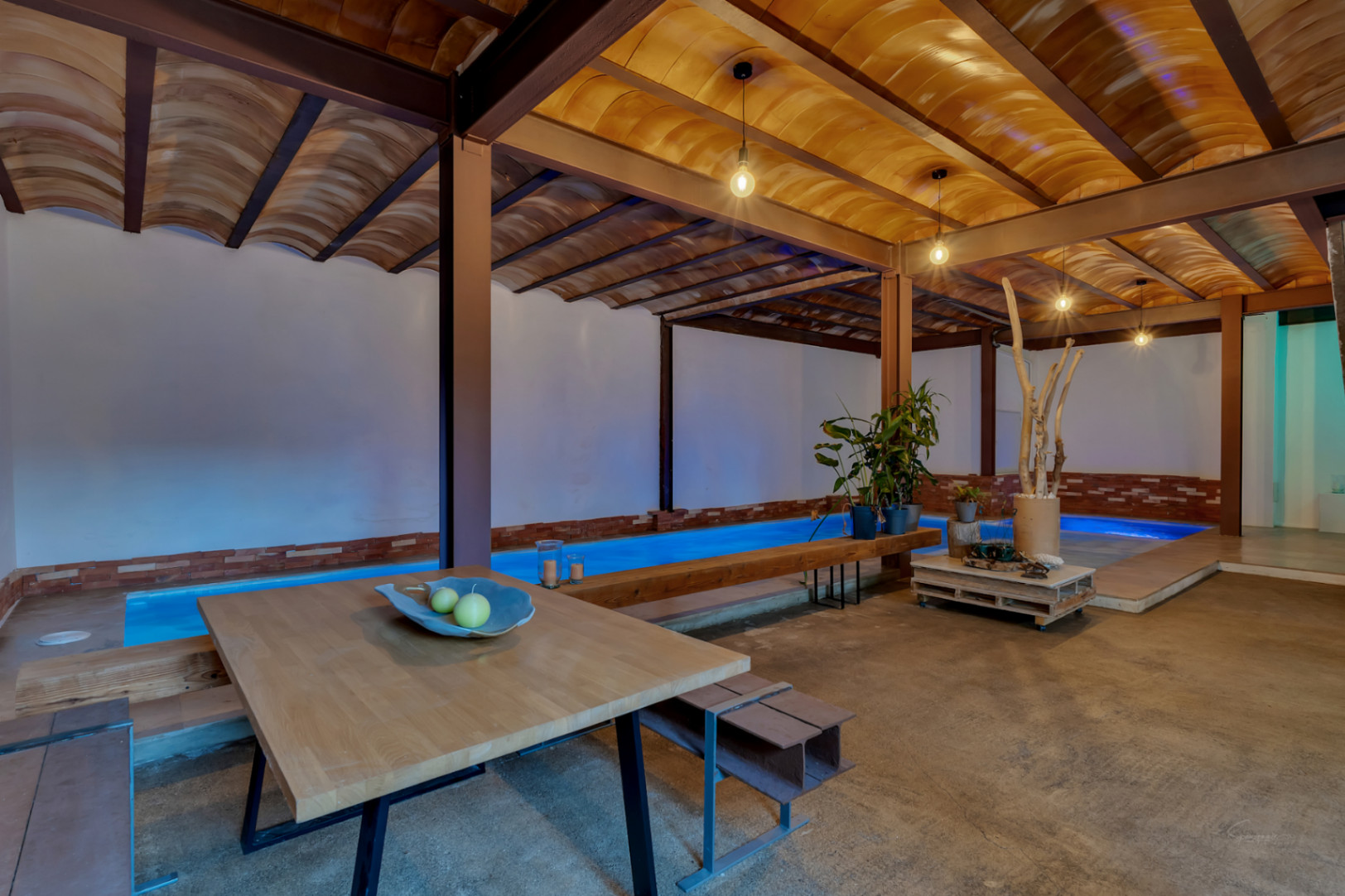 Piscina interior climatizada con iluminación azulada, techos de bovedilla cerámica y vigas de acero. Espacio diáfano con zona de estar y acabados de estilo industrial.