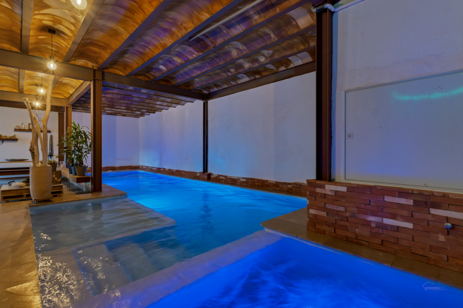 Piscina interior climatizada con iluminación LED azul, techos de bovedilla cerámica y vigas de acero visto que combinan un estilo rústico e industrial.