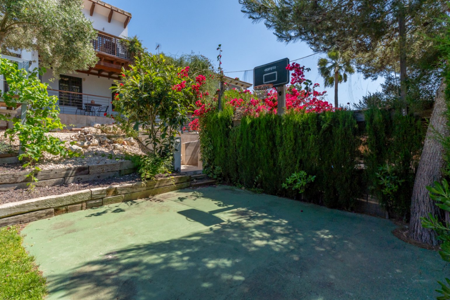Pista de baloncesto privada integrada en el jardín, con superficie verde, canasta y muros de madera rodeados de vegetación mediterránea y buganvillas.