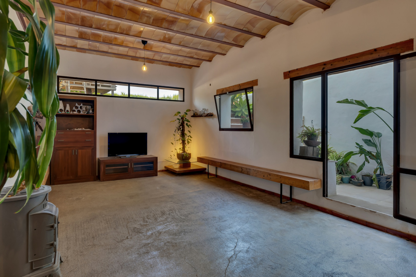 Sala polivalente en semisótano con techos de bovedilla cerámica, suelos de microcemento y acceso directo a un patio interior con vegetación natural.