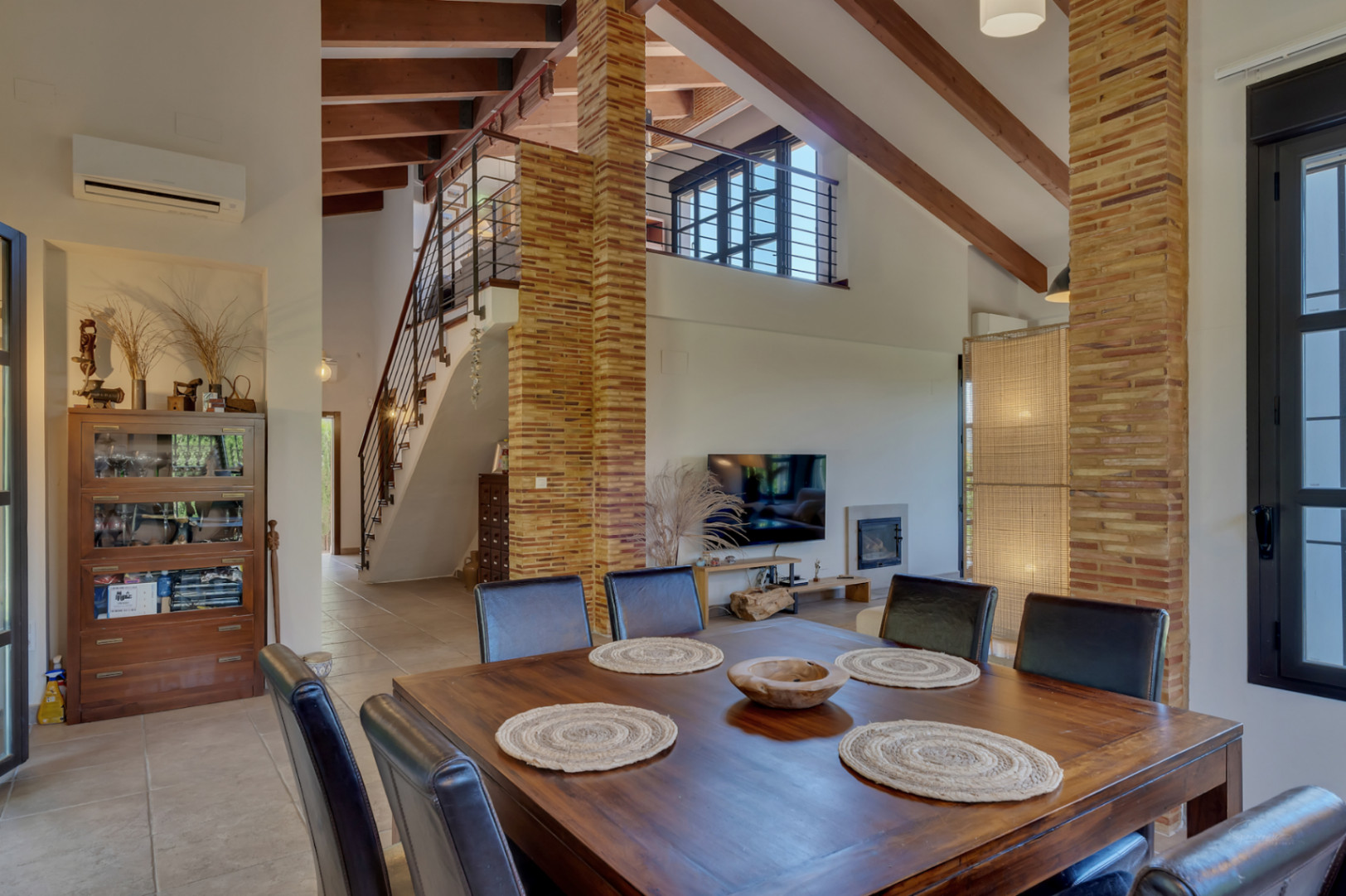 Salón-comedor con techos altos y vigas de madera, destacando sus pilares de ladrillo visto y la escalera que da acceso al dormitorio superior abierto.