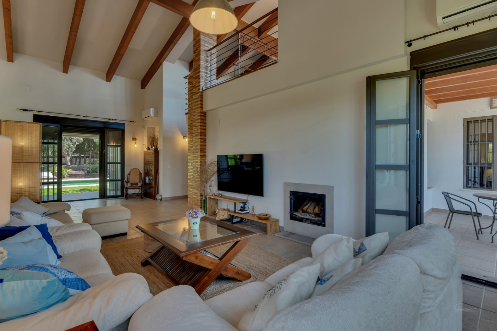 Salón de techos altos con vigas de madera expuestas, chimenea integrada y acceso directo a la terraza y jardín a través de amplios ventanales.