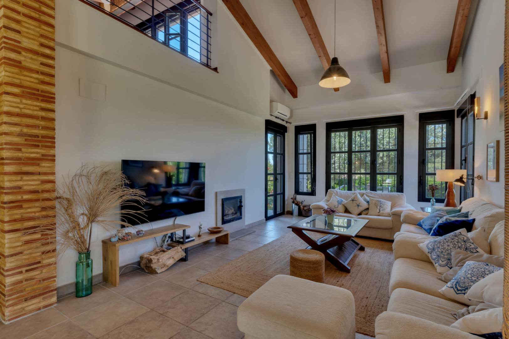 Salón de techos altos con vigas de madera expuestas, chimenea integrada y grandes ventanales que aportan luz natural y acceso directo al jardín.