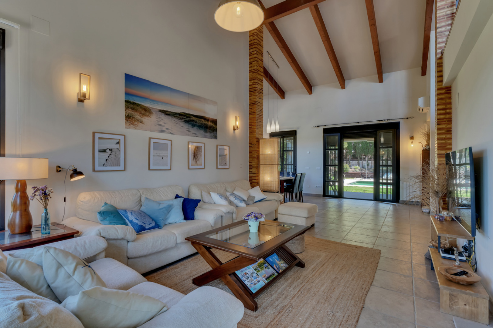 Salón de techos altos con vigas de madera expuestas, pilares de ladrillo visto y acceso directo al jardín y la piscina a través de amplios ventanales.