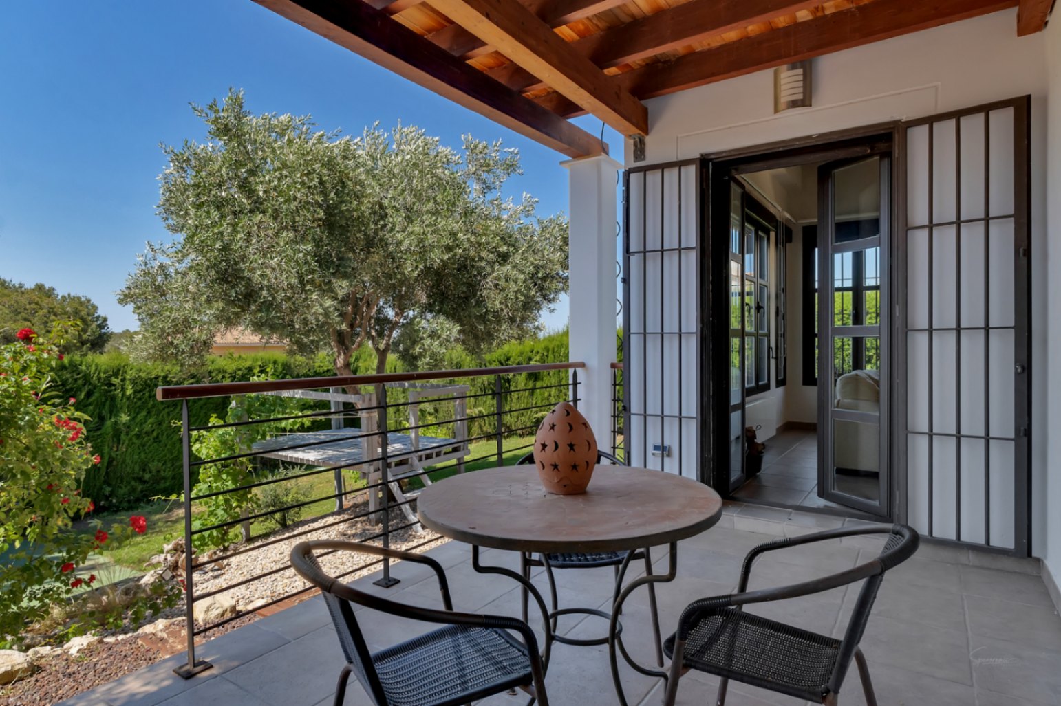 Terraza cubierta con vigas de madera, suelo cerámico y mobiliario de forja, ofreciendo vistas despejadas al jardín mediterráneo y al olivo central.