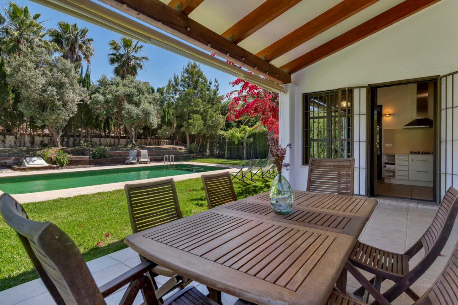 Terraza cubierta con vigas de madera y comedor exterior, con vistas directas al jardín mediterráneo y la piscina rectangular rodeada de césped.