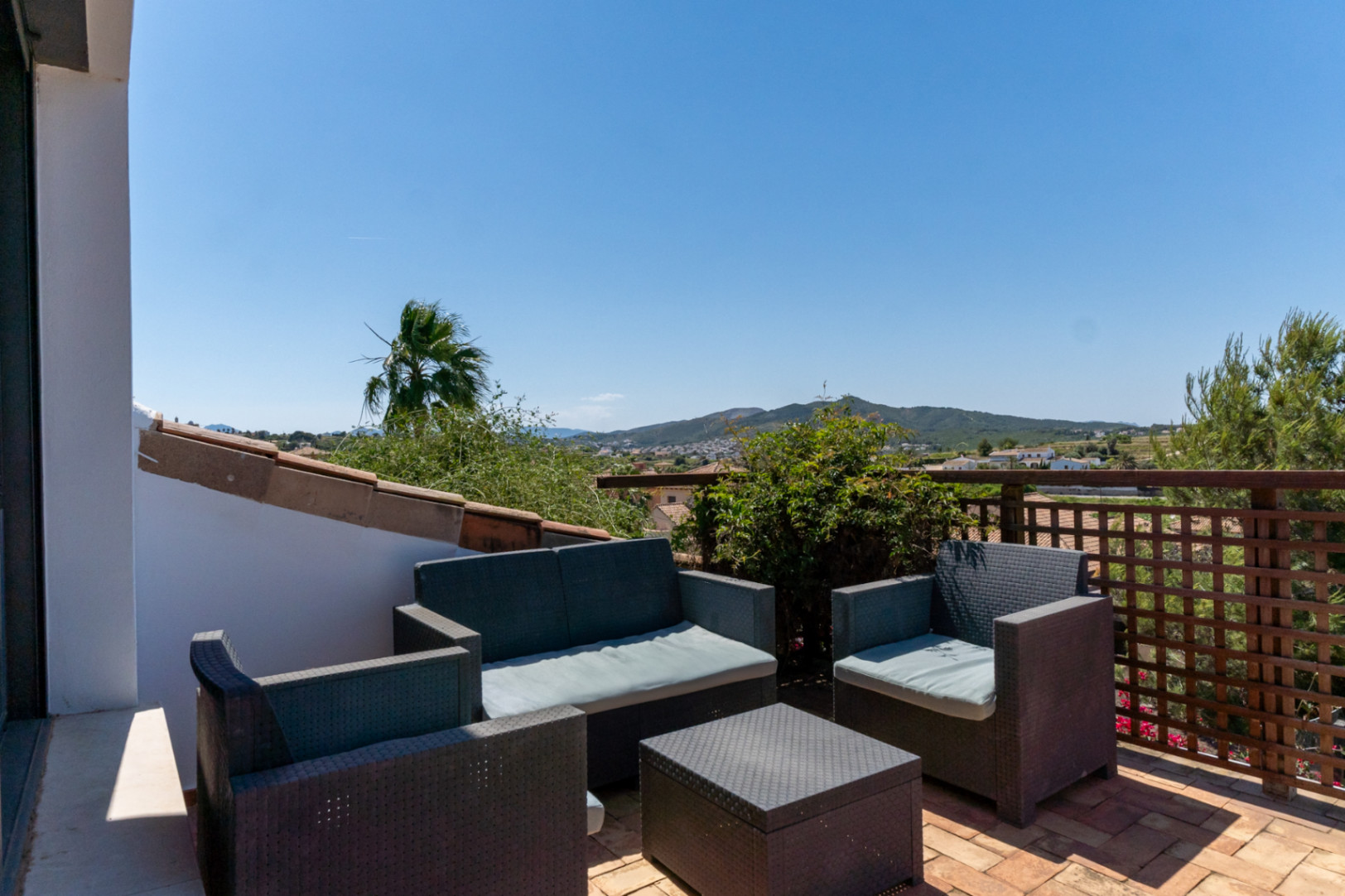 Terraza superior con mobiliario de exterior en ratán sintético y suelos de barro, ofreciendo vistas despejadas al valle y las montañas de Jávea.
