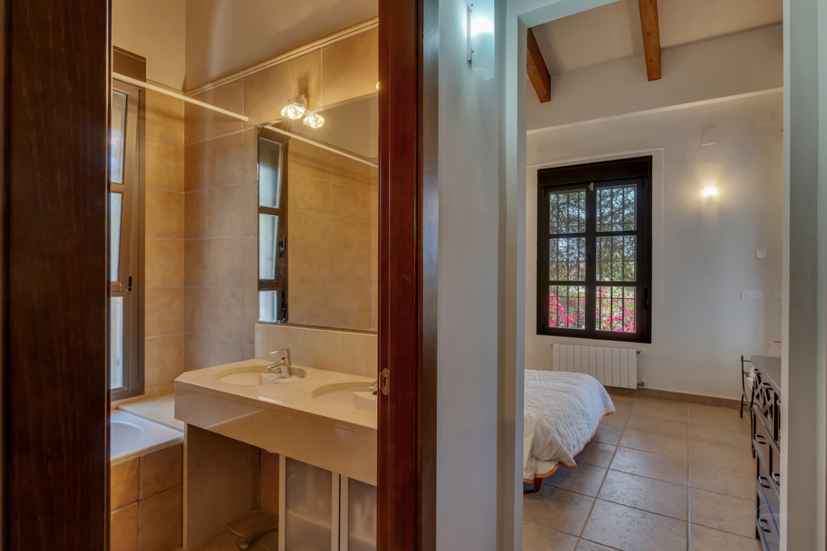 Vista desde el pasillo hacia el baño con lavabo doble de piedra y un dormitorio luminoso con vigas de madera vistas y suelos cerámicos.