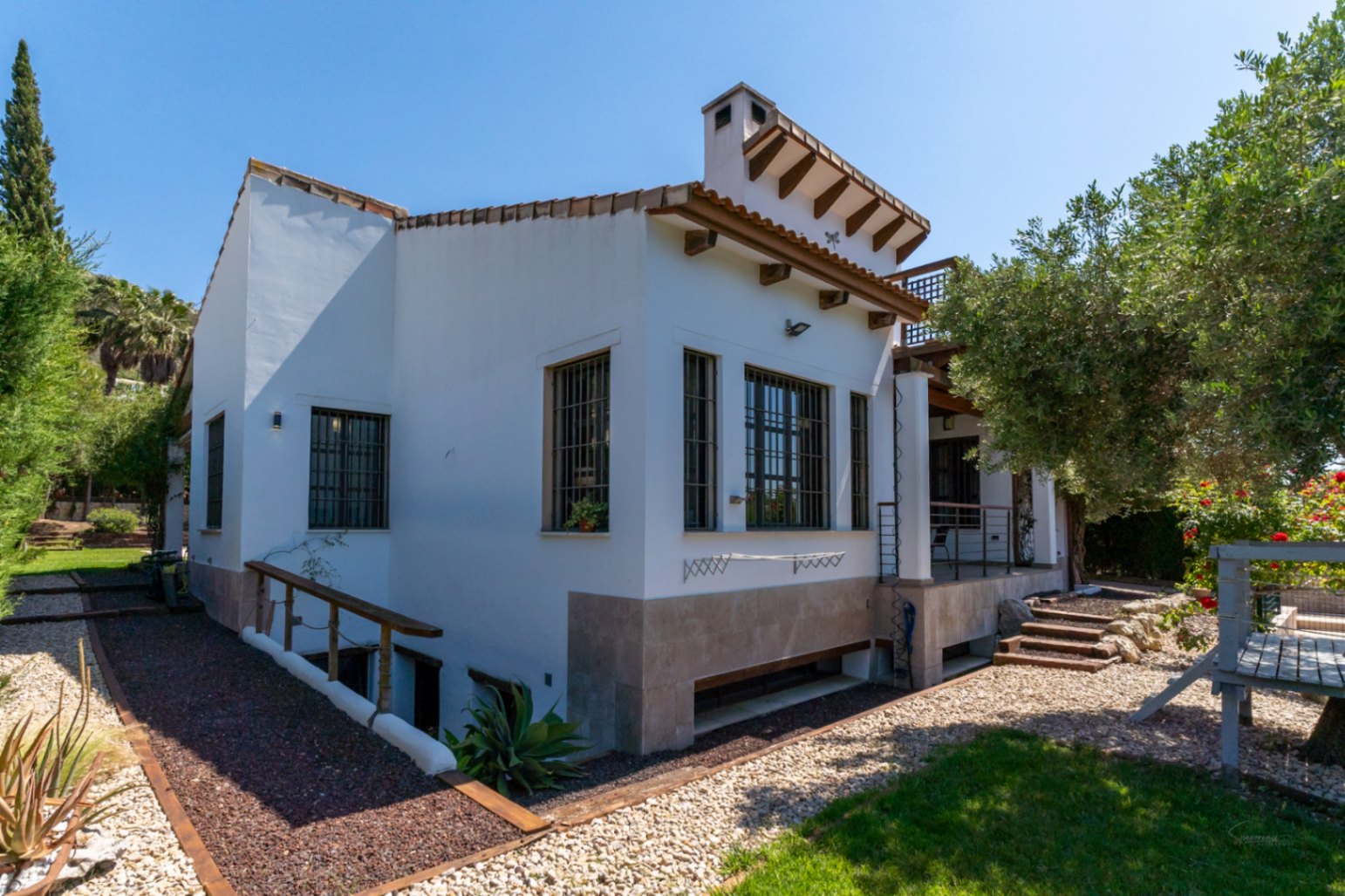 Vista exterior de la villa con fachada blanca, vigas de madera y zócalo de piedra. El jardín cuenta con senderos de grava y acceso descendente al semisótano.