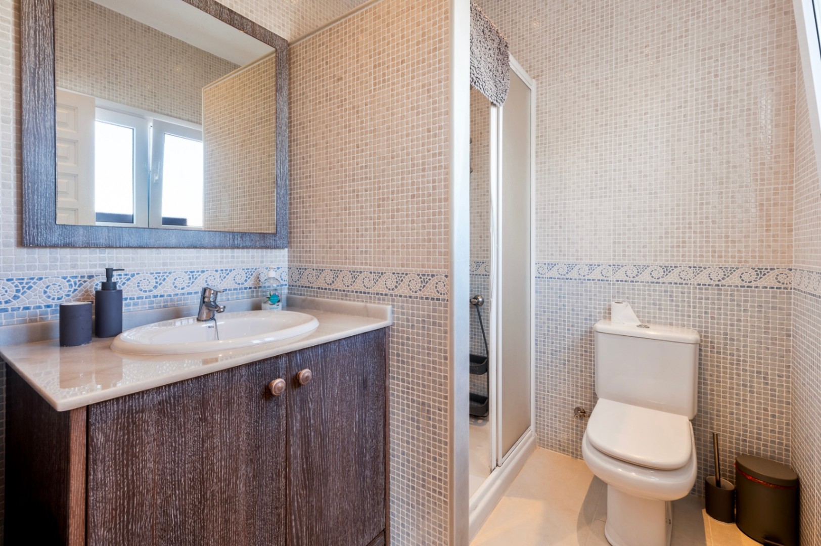 Baño con paredes de mosaico en tonos crema, cenefa decorativa azul, mueble de madera oscura con encimera de mármol y ducha integrada.