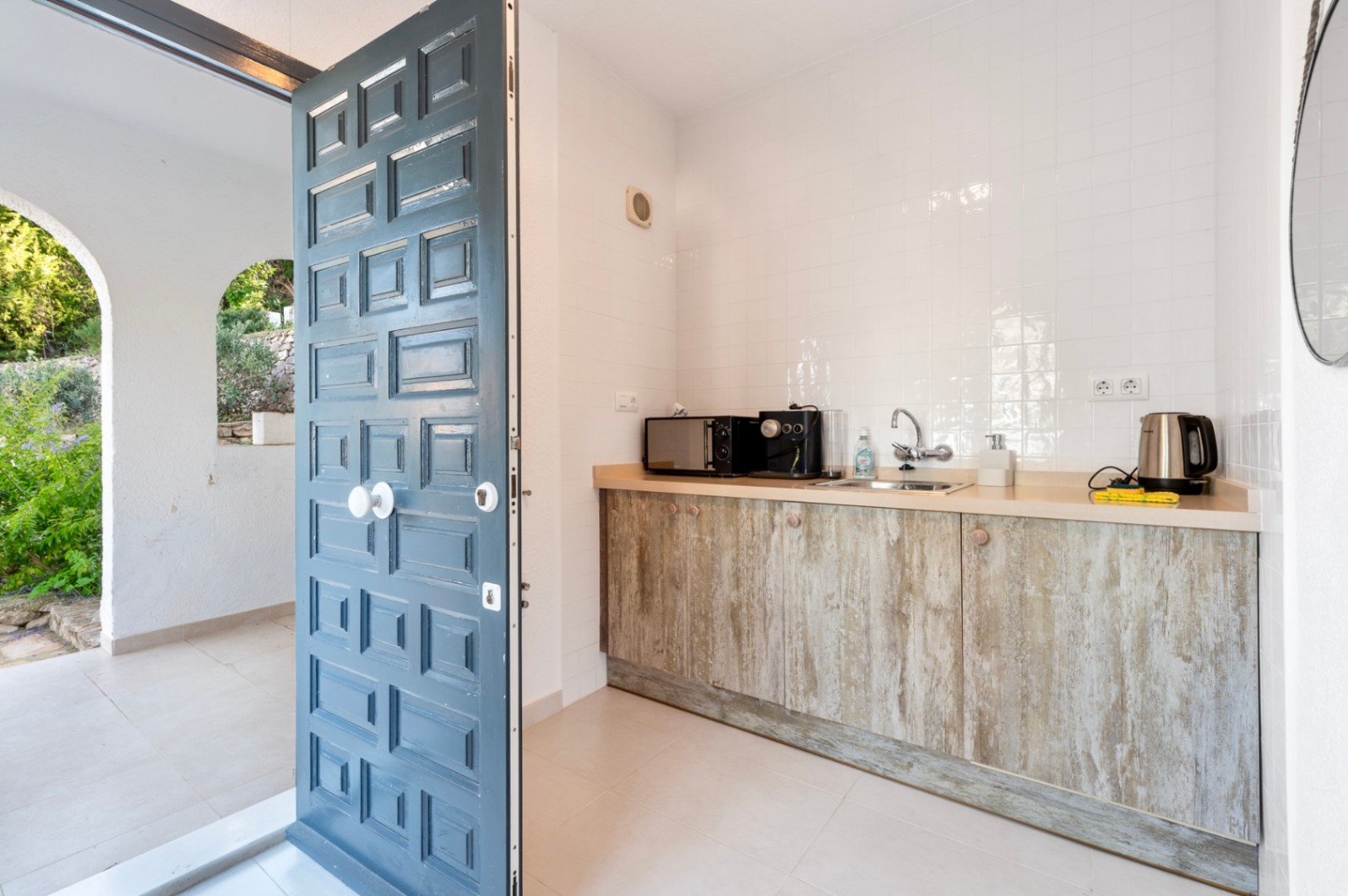 Cocina auxiliar compacta con armarios de efecto madera rústica y encimera clara, situada junto a una puerta de cuarterones azul que da acceso al exterior.