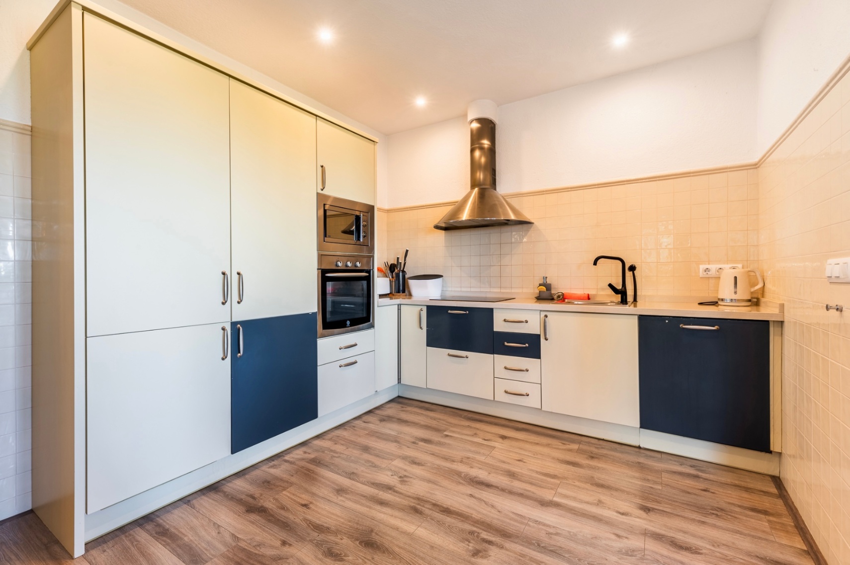 Cocina moderna reformada con armarios bicolores en blanco y azul, encimera clara, electrodomésticos de acero inoxidable y suelo de tarima resistente.