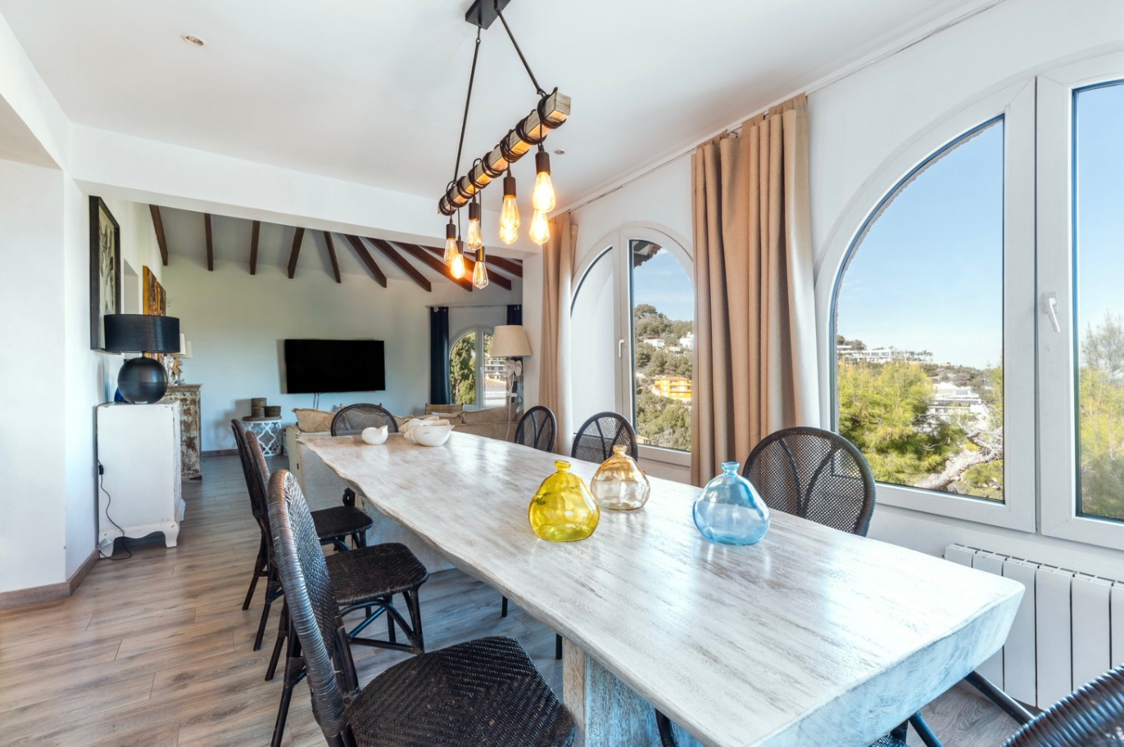 Comedor luminoso con mesa de madera rústica, sillas de mimbre negro y amplios ventanales de arco con vistas. Destaca la lámpara de estilo industrial y el techo con vigas de madera.